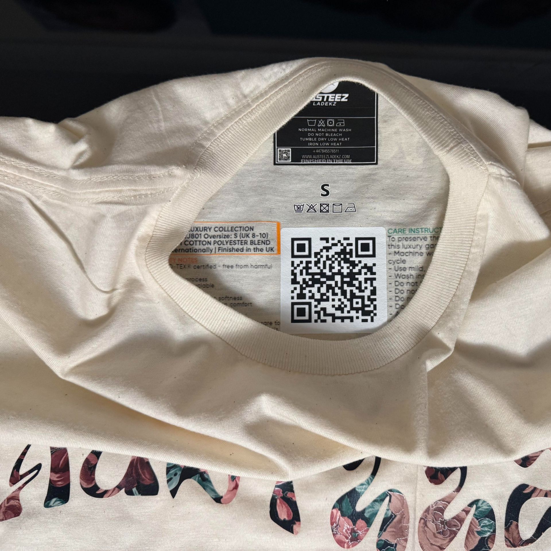 Cream-colored t-shirt, showing the inside neck with tags. QR code and size "S" label are visible.