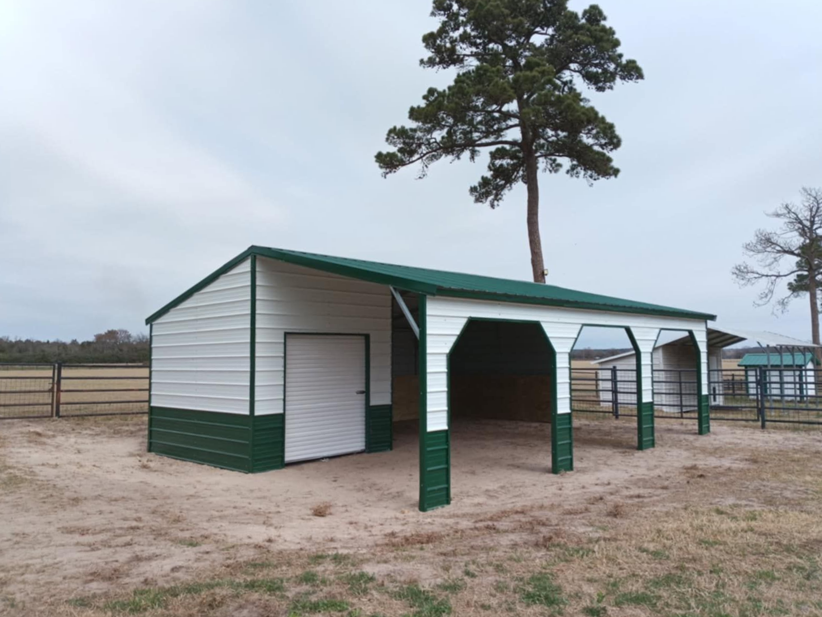 White barn-style shelter with green trim in a fenced dirt field, with a tall tree behind it.