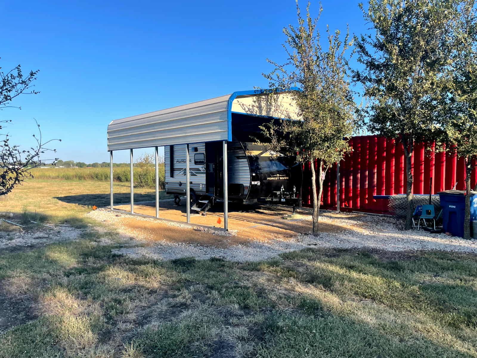 Metal carport beside a red container on a grassy rural lot under a clear blue sky
