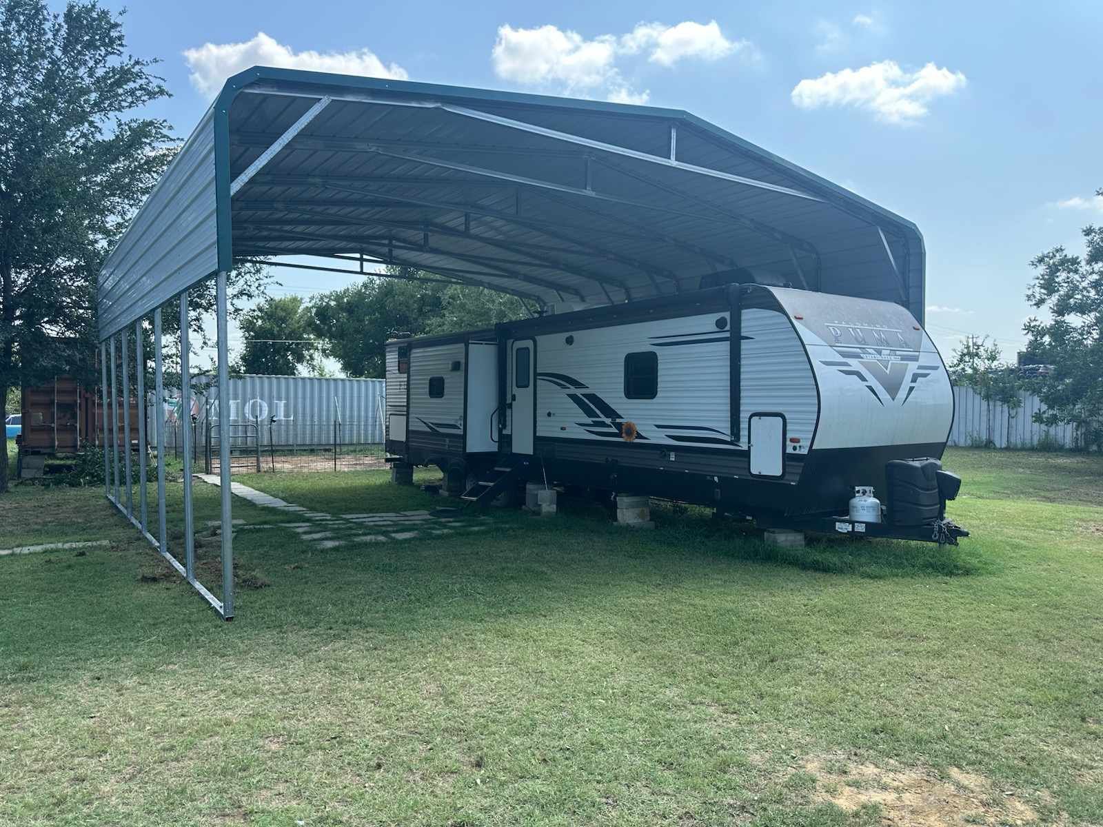 White RV parked under a metal carport on a grassy lot, with trees and a fence in the background