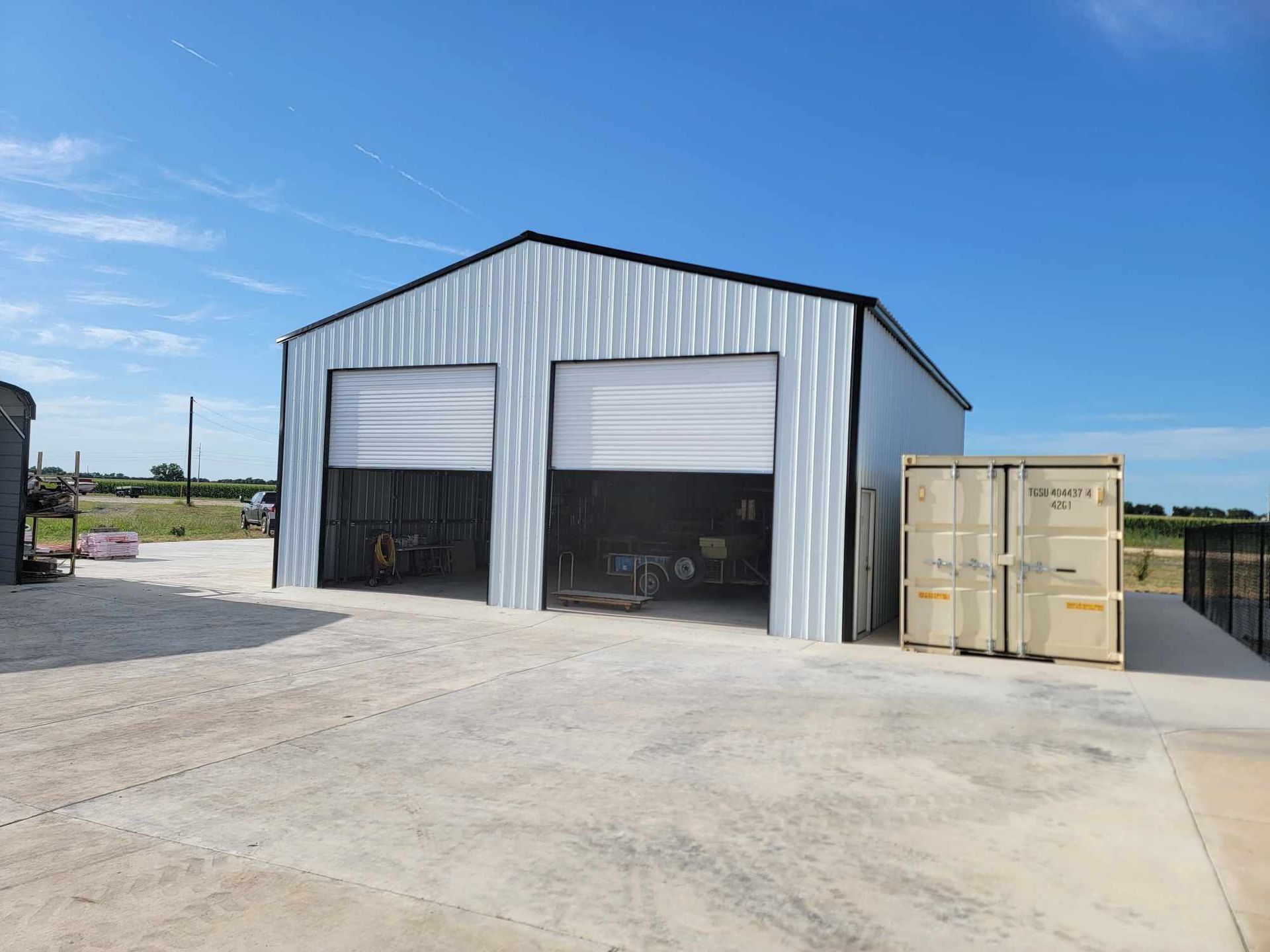 Metal warehouse with two open garage bays and tan storage containers on a concrete lot under a blue sky