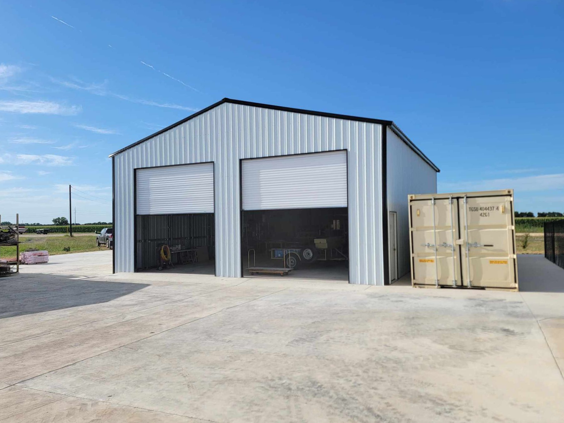 Metal warehouse with two open bays on a concrete lot beside stacked shipping containers under a blue sky
