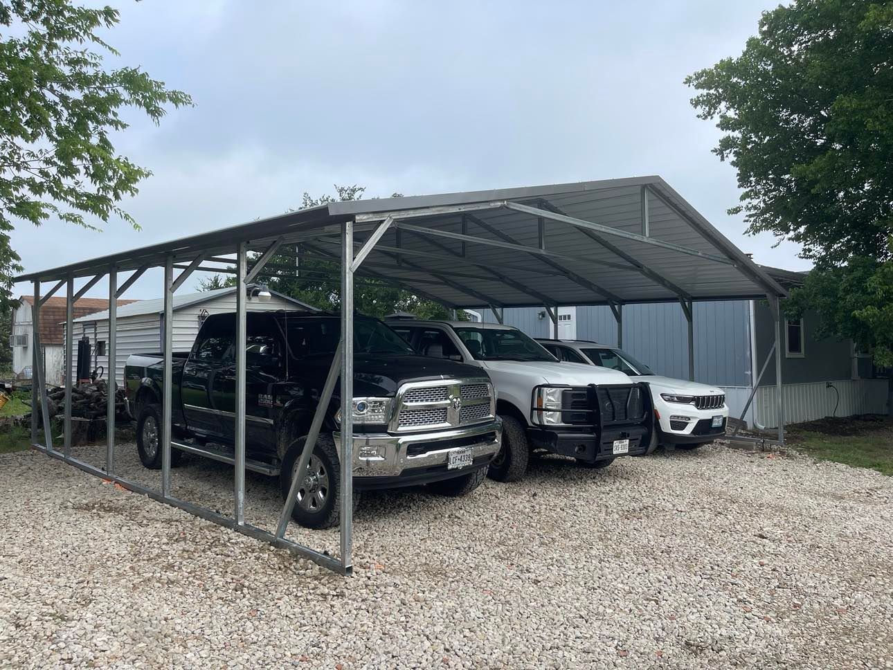 Several cars parked under a metal carport on a gravel driveway, flanked by trees and a building.