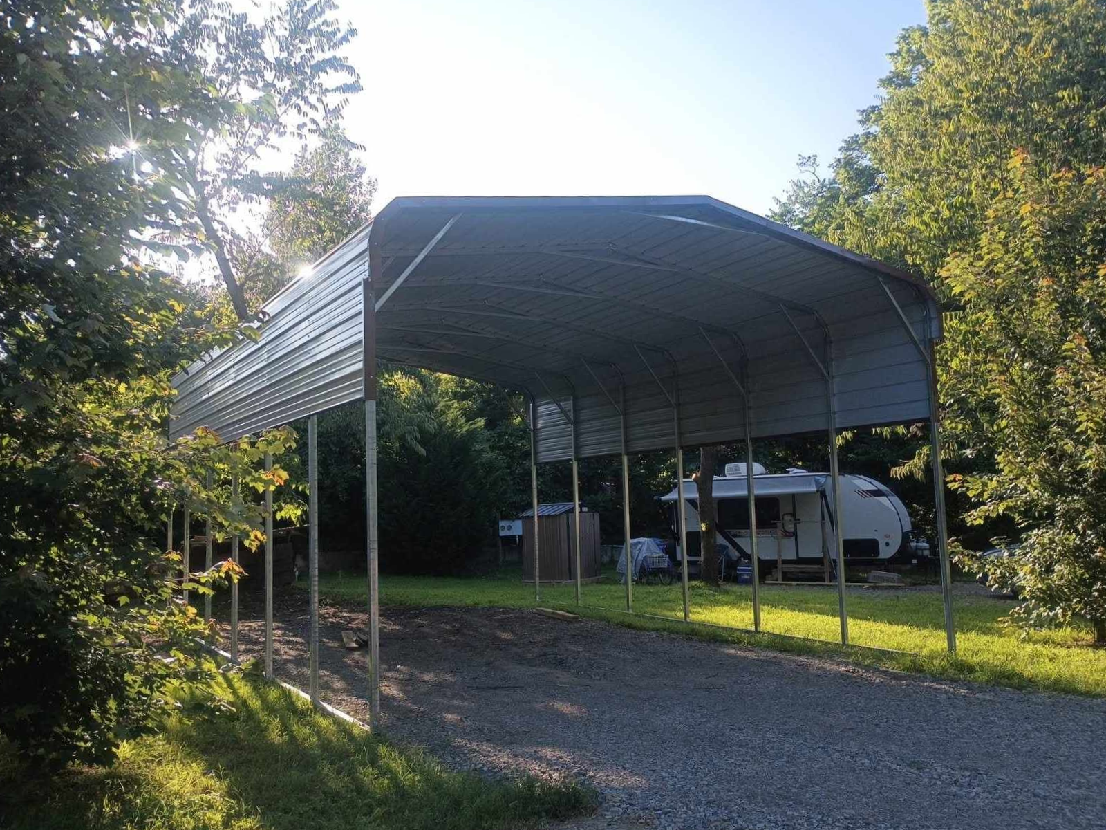 Long metal carport in a gravel driveway, with a white van parked underneath and trees in the background