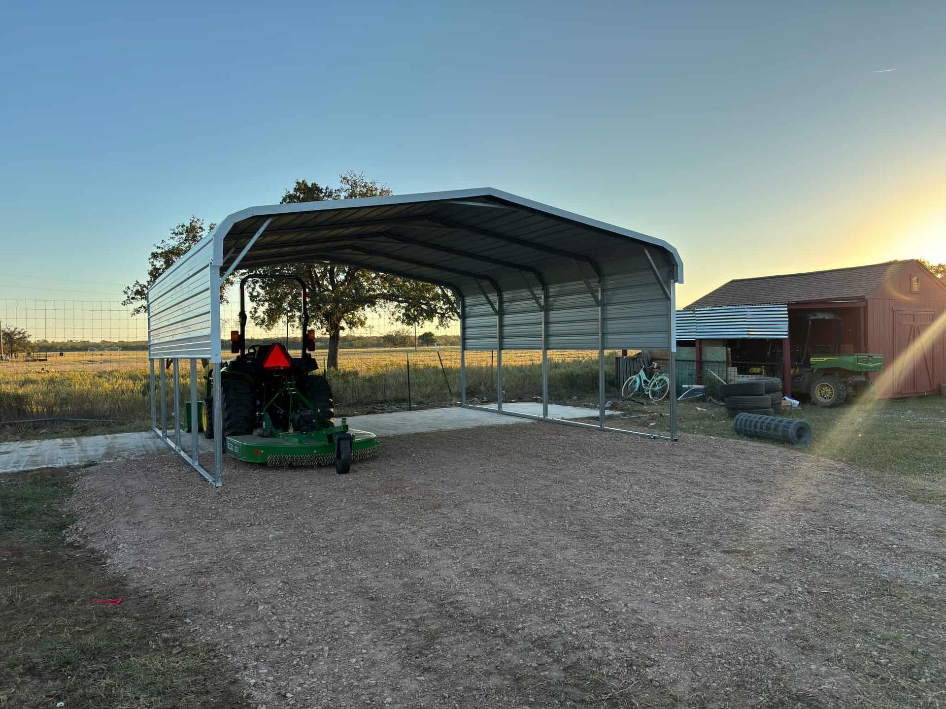 Open-sided metal shed on a gravel lot at sunset, with a tractor inside and trees in the background