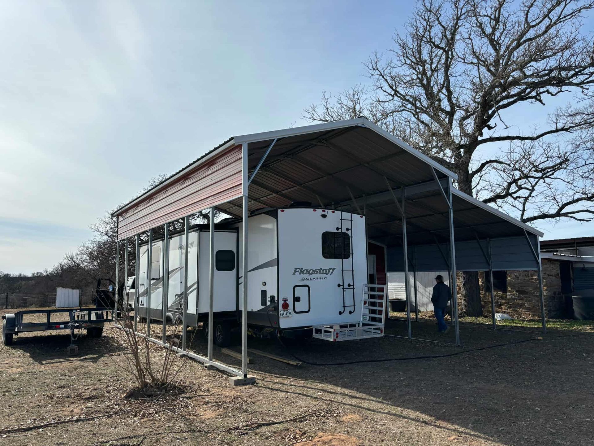 Camper trailer parked under a metal carport in a rural yard, with trees and a trailer nearby.
