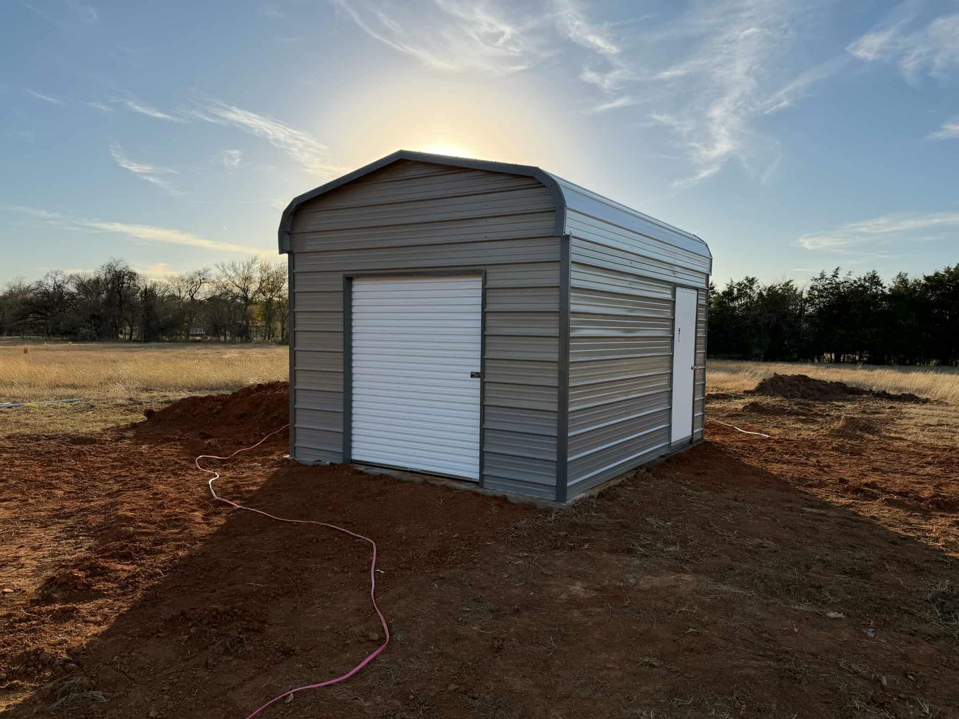 Gray metal storage shed in a dirt field at sunset, with a roll-up door and side window.