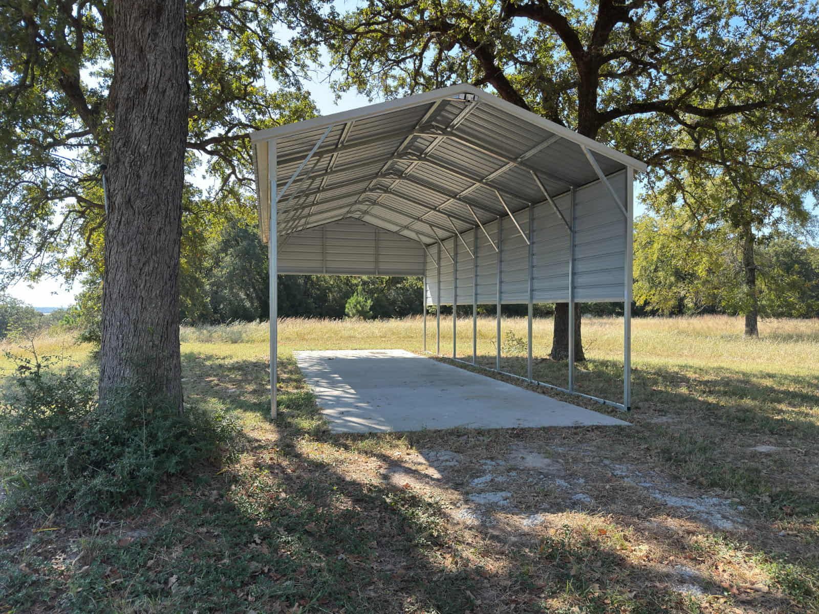 Open-sided metal shelter with a concrete floor in a grassy, tree-filled park area.