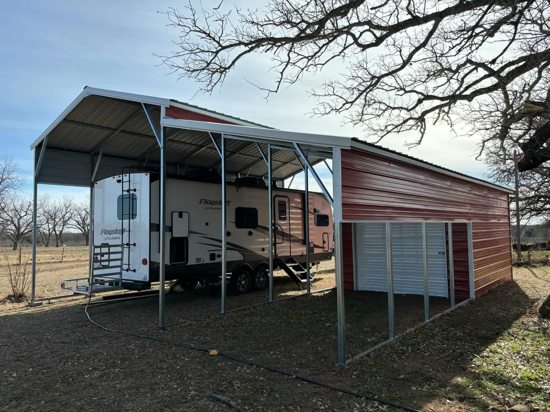 Camper trailer under a metal carport beside a red shed in a rural field