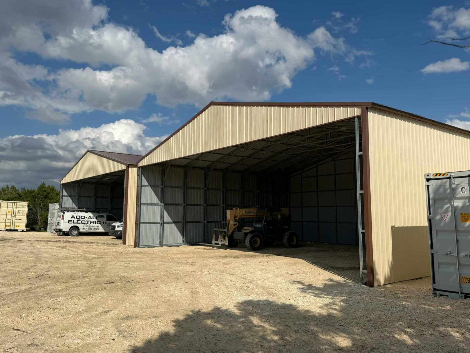 Open-sided beige storage shed with vehicles inside under a partly cloudy sky