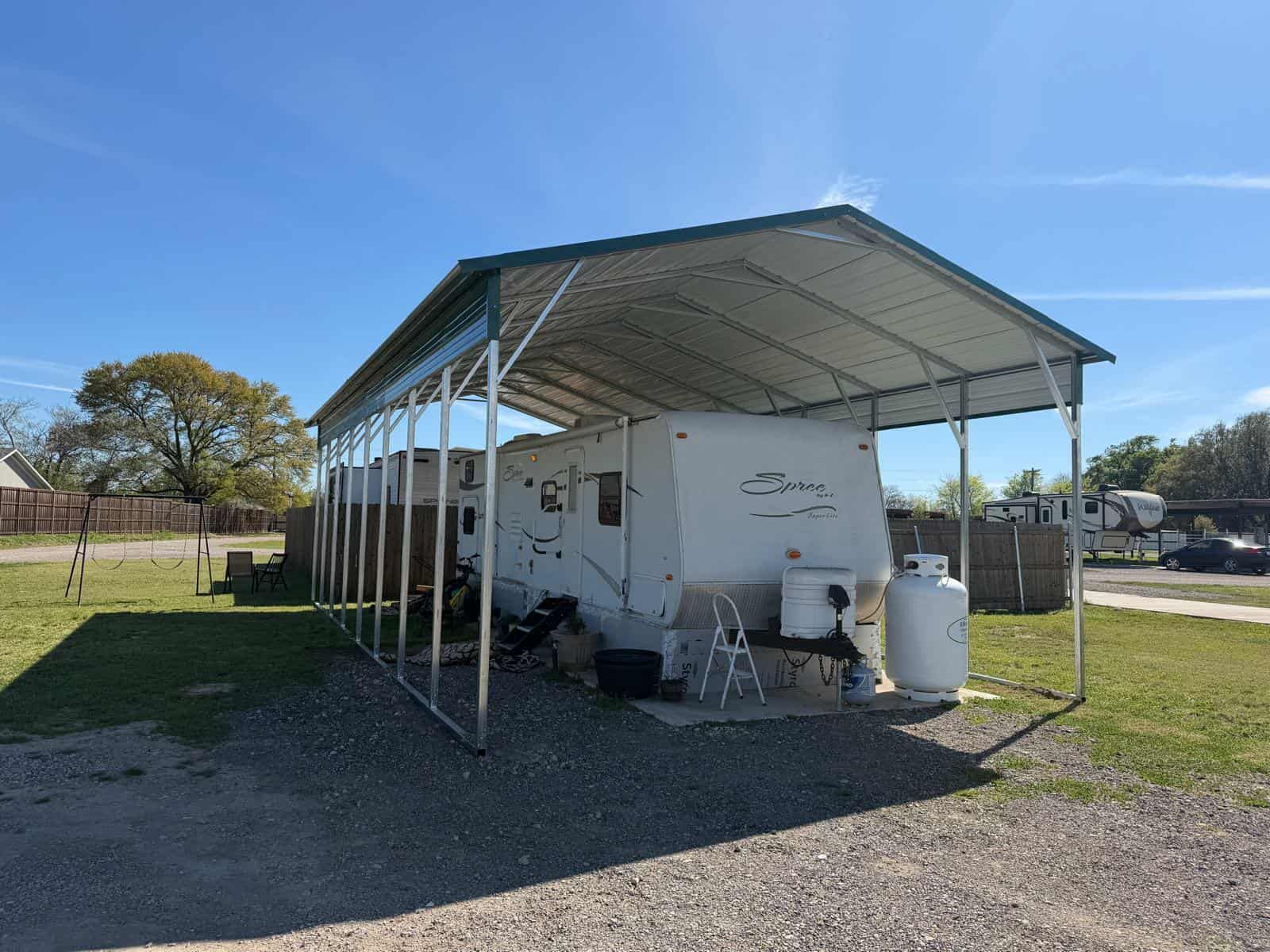 Small white outbuilding with metal awning in a grassy yard under a clear blue sky