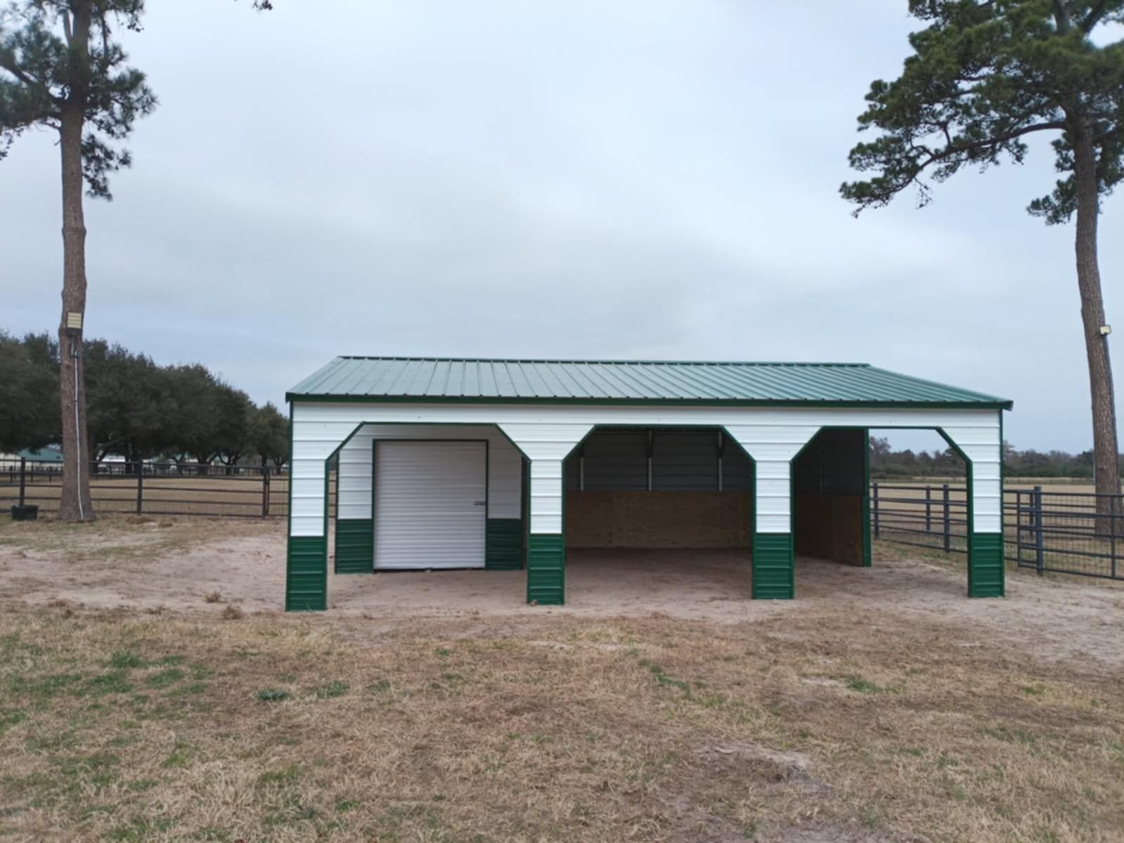 Green-roofed pavilion with open bays in a sandy park clearing, framed by tall pine trees