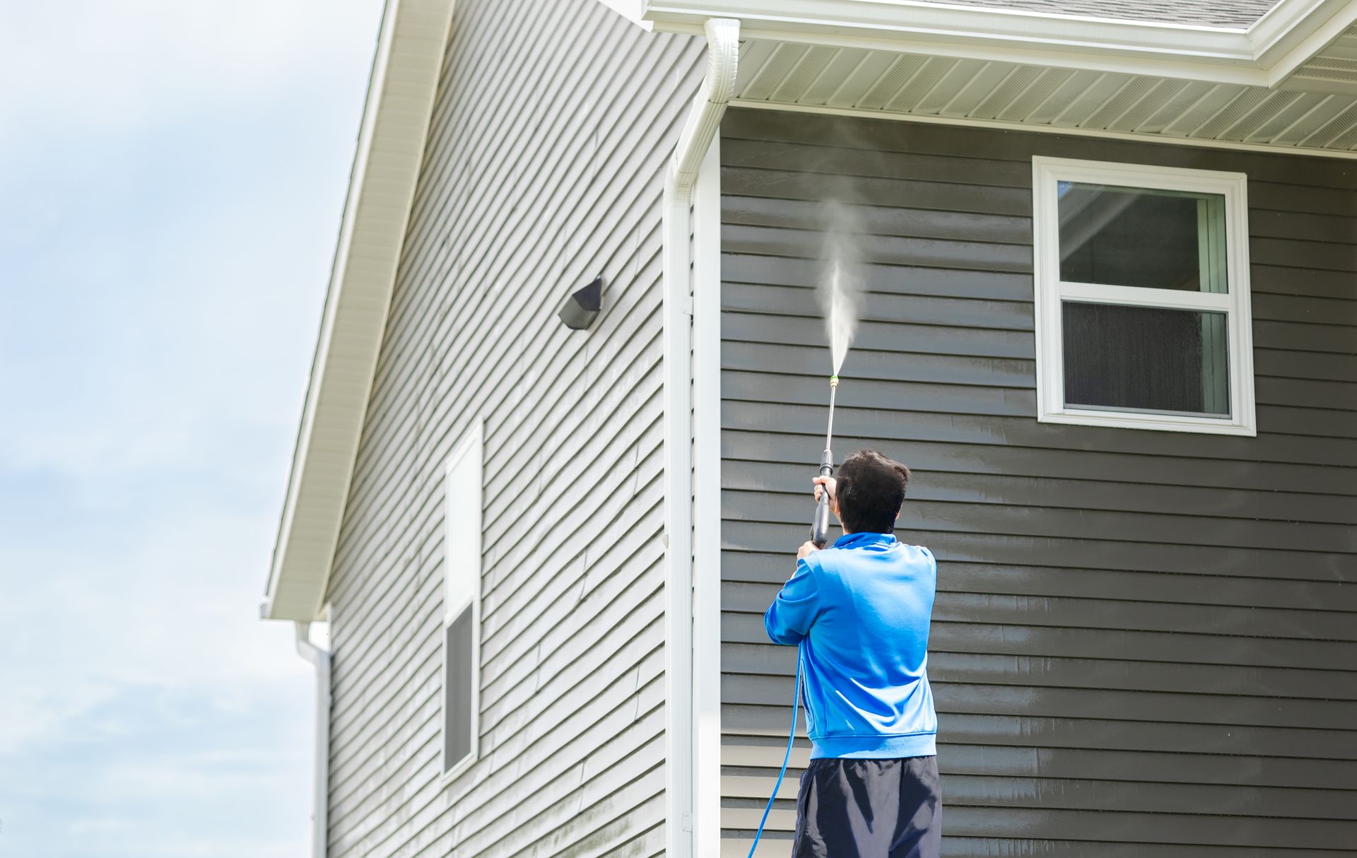 Man in blue jacket cleans dusk and dirt from exterior siding and under roof with a high-pressure nozzle spray with water soap cleaner