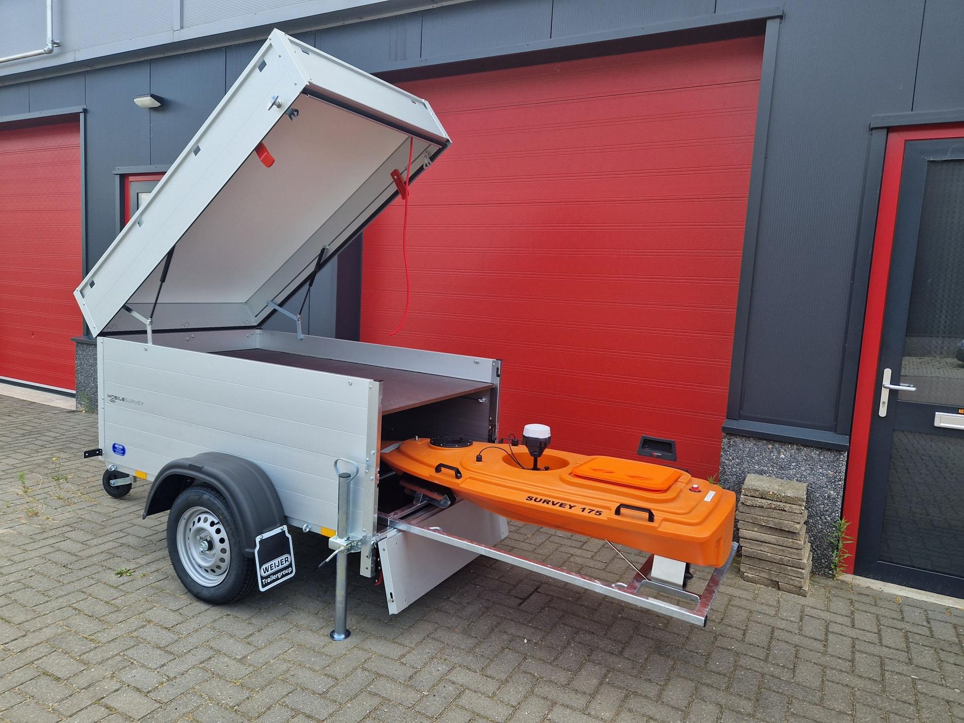 Voor een rode garagedeur staat een aanhangwagen met daarin een oranje kajak geparkeerd.