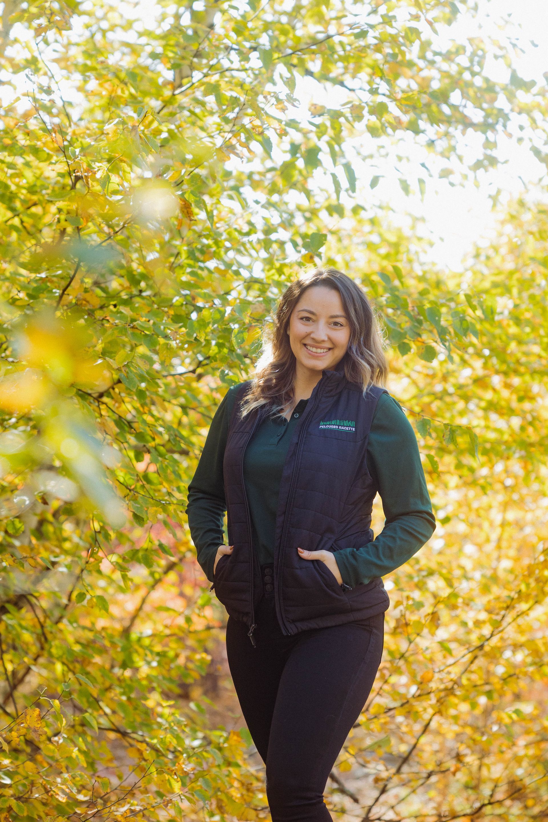 Une femme vêtue d’un gilet noir se tient devant un arbre.