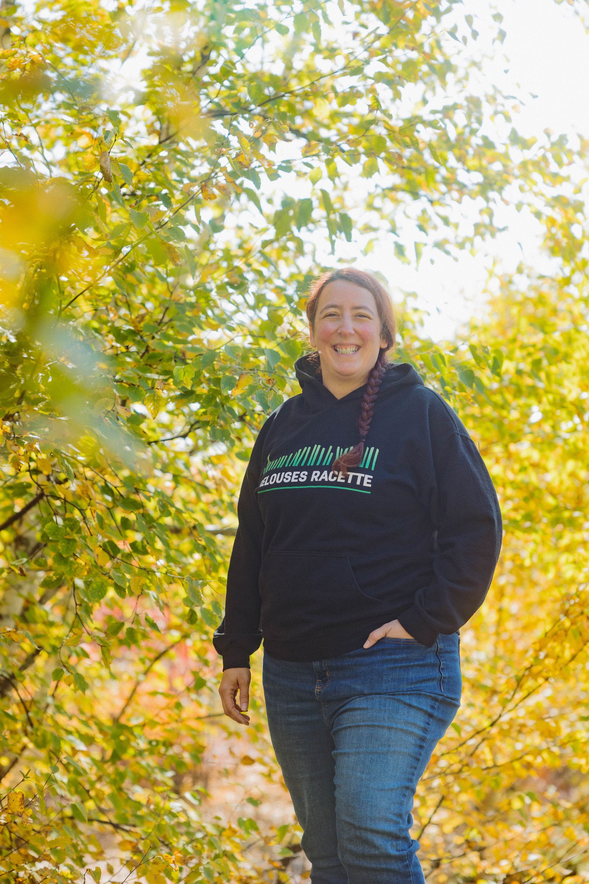 Une femme portant un sweat à capuche noir et un jean se tient devant un arbre.