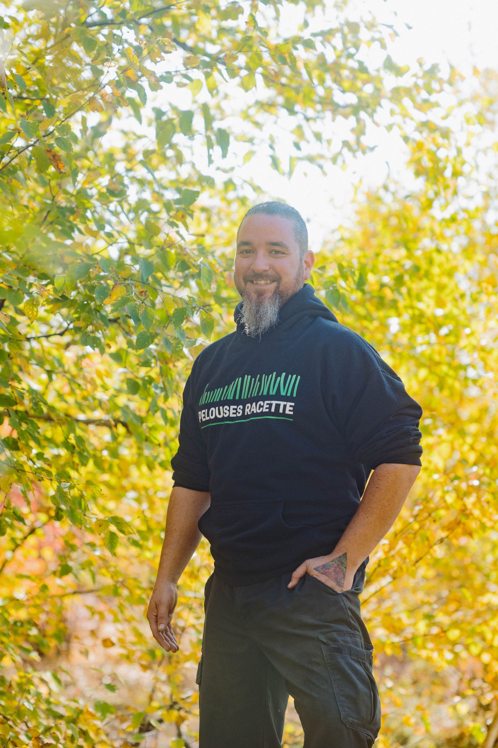 Un homme barbu se tient dans les bois et porte un sweat à capuche noir.