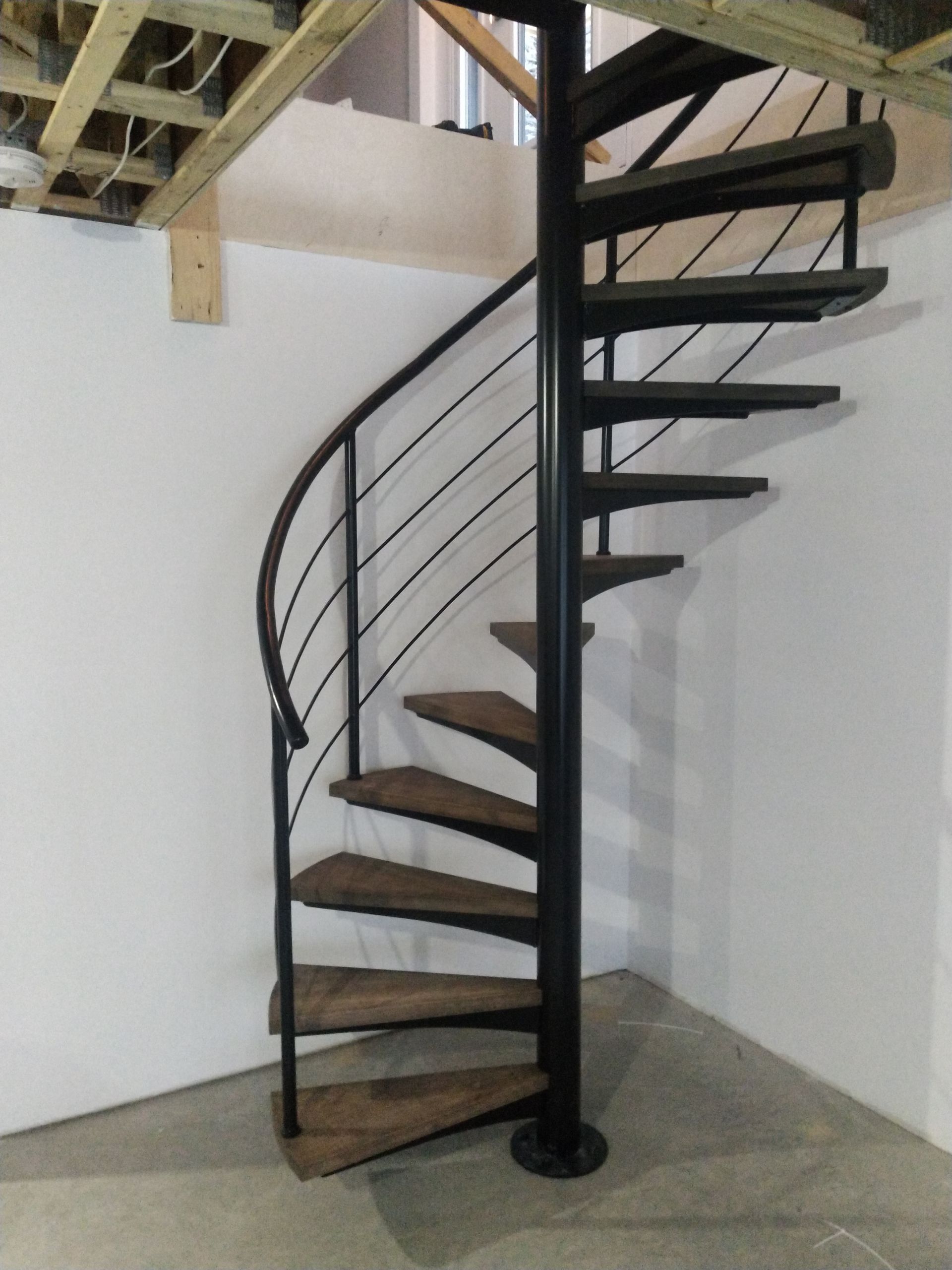 Escalier en colimaçon avec structure en métal noir et marches en bois, contre un mur blanc.