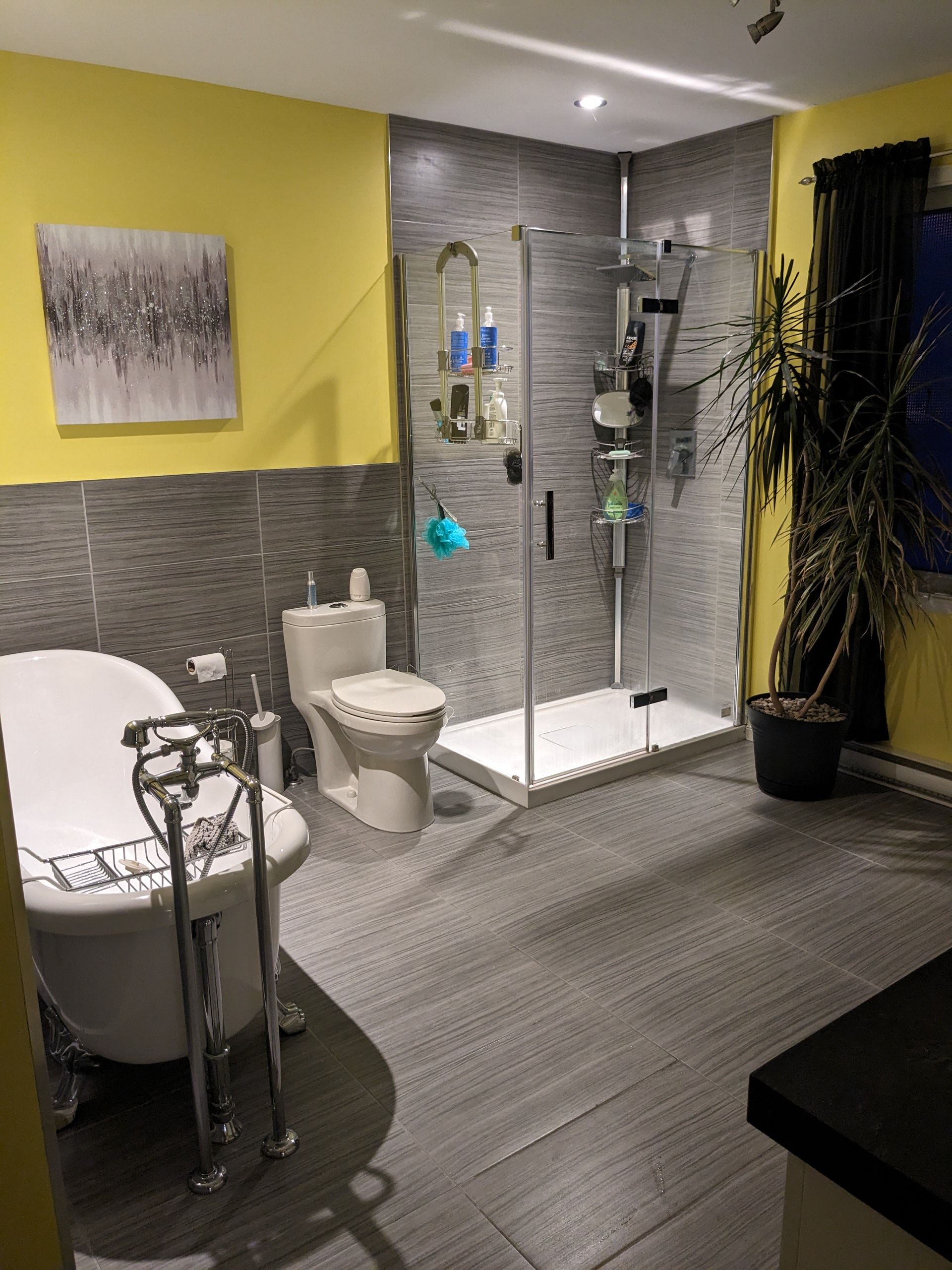 Salle de bain avec murs jaunes, sols/murs carrelés gris, baignoire sur pieds, douche en verre et toilettes.