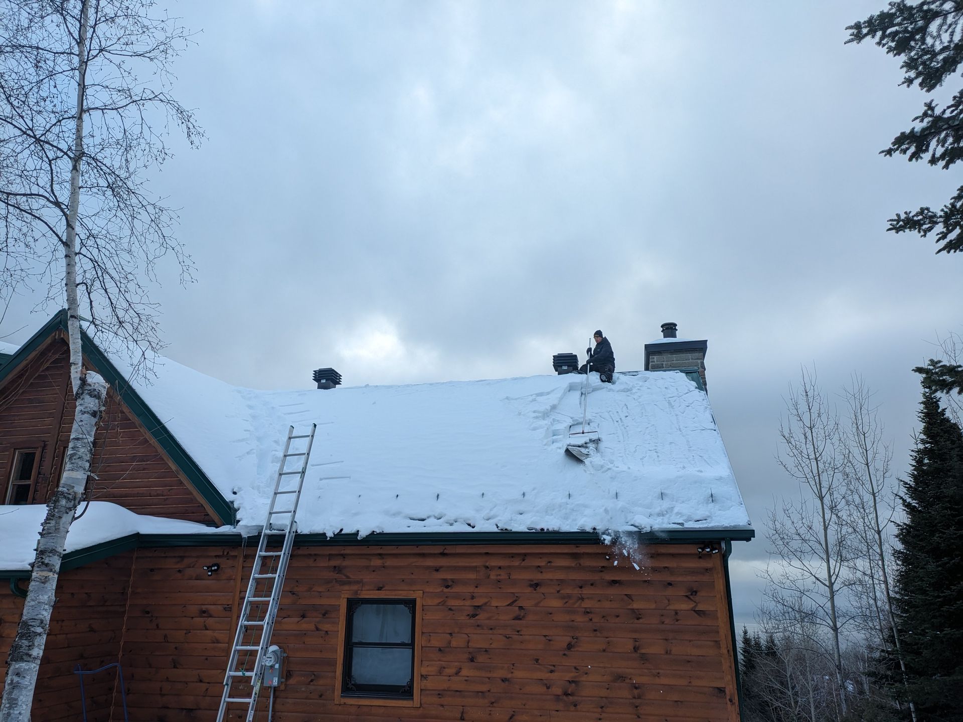 Personne sur un toit enneigé déblayant la neige d'une cheminée à côté d'une échelle, maison en bois, ciel nuageux.