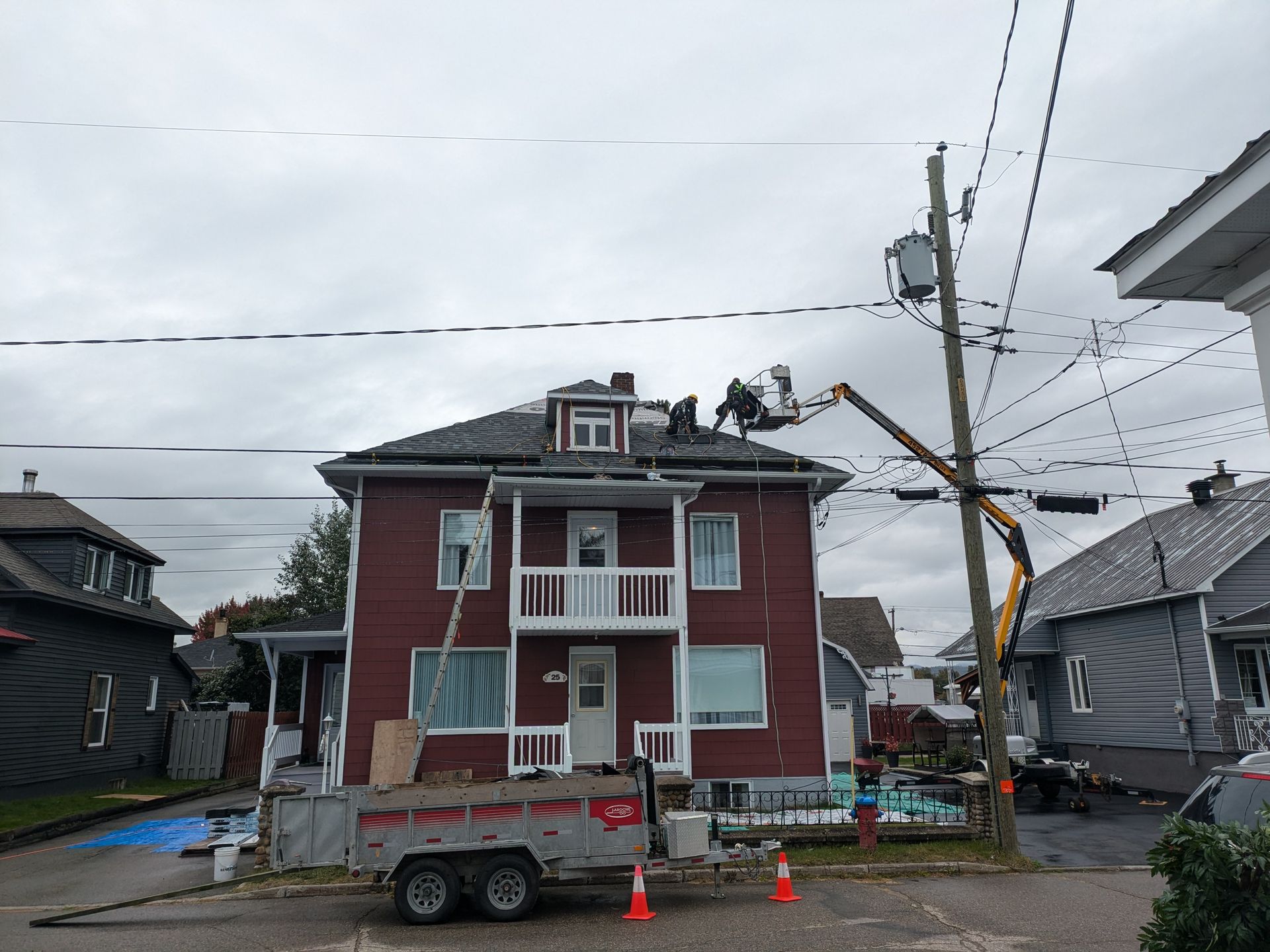 Des ouvriers sur un toit près de lignes électriques, aidés par un ascenseur. Maison rouge au toit sombre, ciel gris.