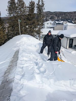 Deux personnes pelletent la neige d'un toit par une journée ensoleillée, avec des maisons couvertes de neige en arrière-plan.