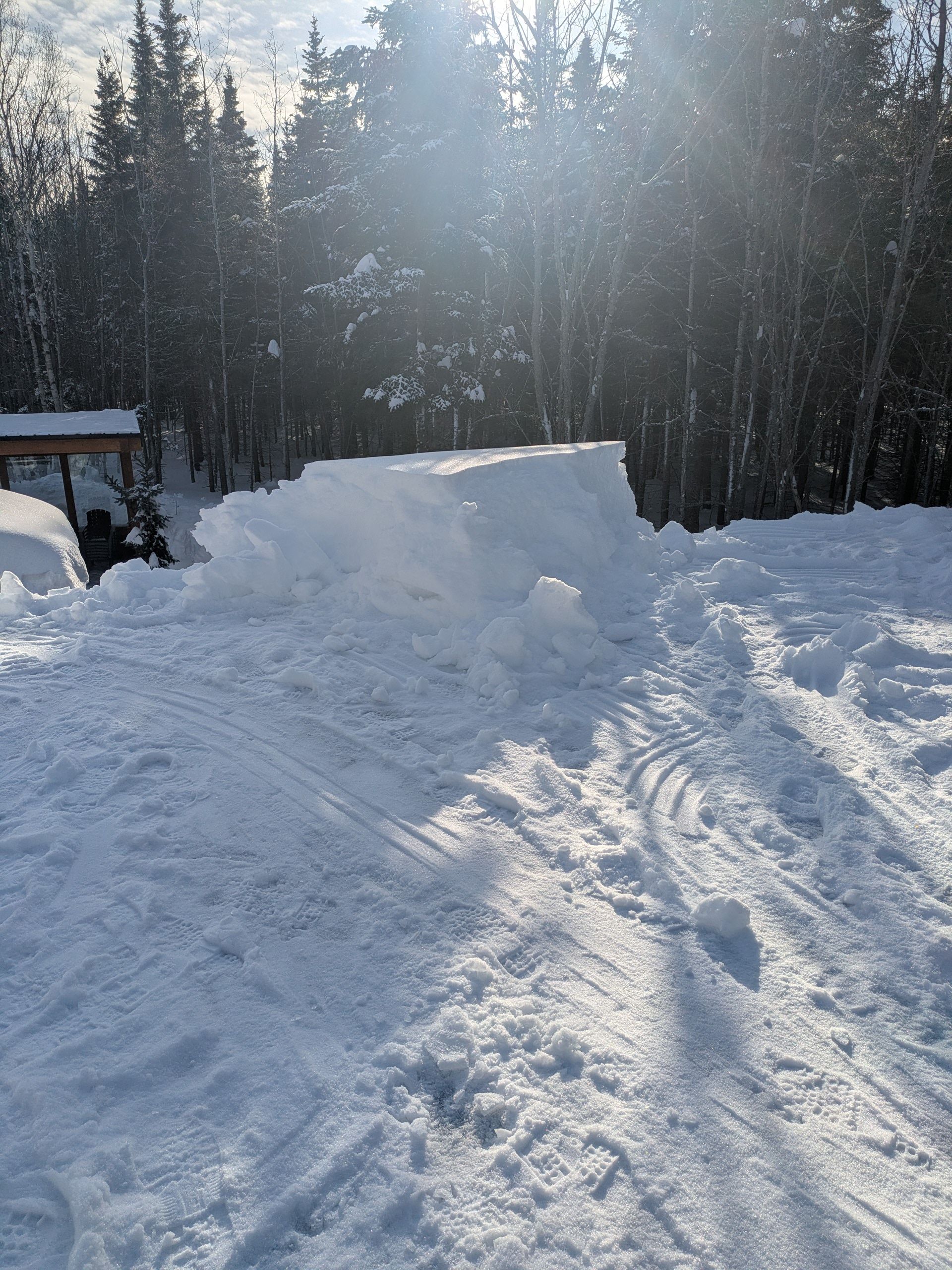 Tas de neige dans une cour enneigée. Arbres et soleil en arrière-plan.