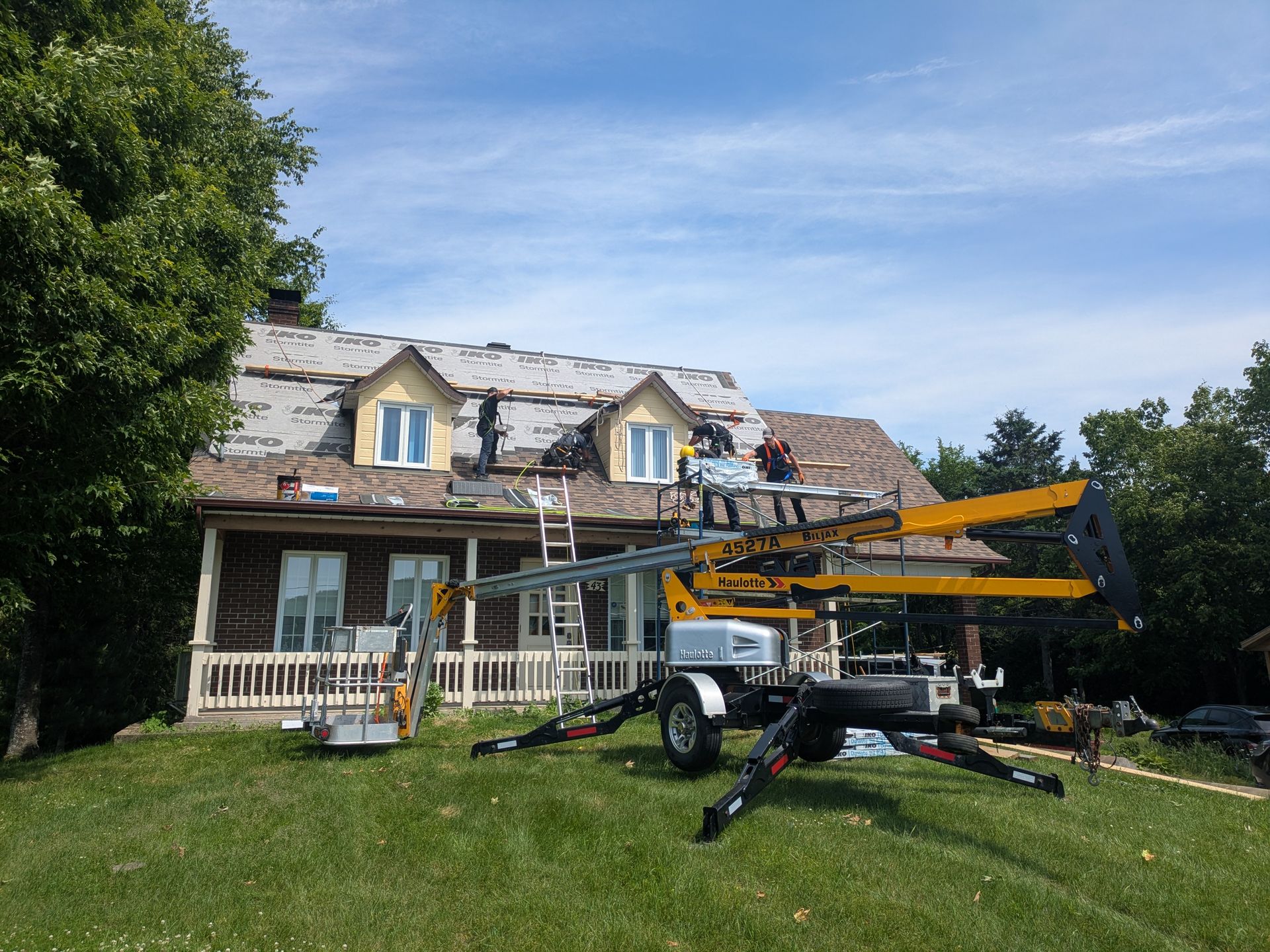 Des couvreurs utilisent une nacelle élévatrice pour remplacer un toit en bardeaux sur une maison à deux étages ; journée ensoleillée.
