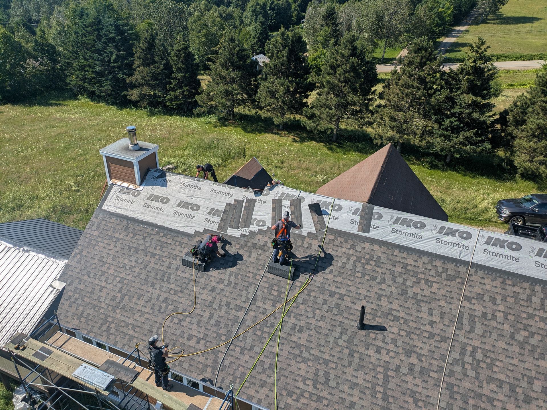 Couvreurs installant des bardeaux sur le toit d'une maison ; champ vert et arbres en arrière-plan.