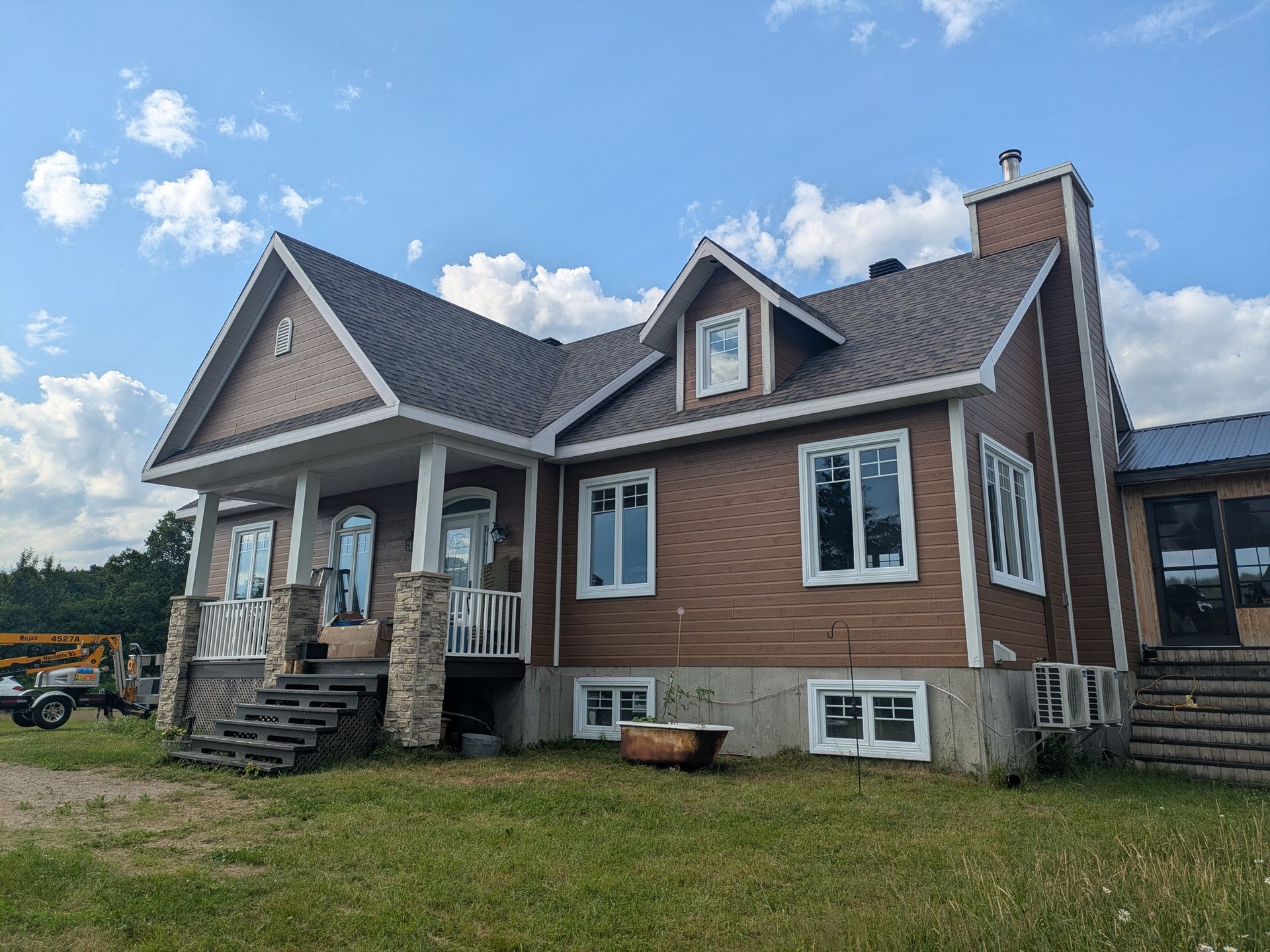 Maison brune avec porche, cheminée et fenêtres à cadre blanc sur un terrain herbeux vert sous un ciel nuageux.
