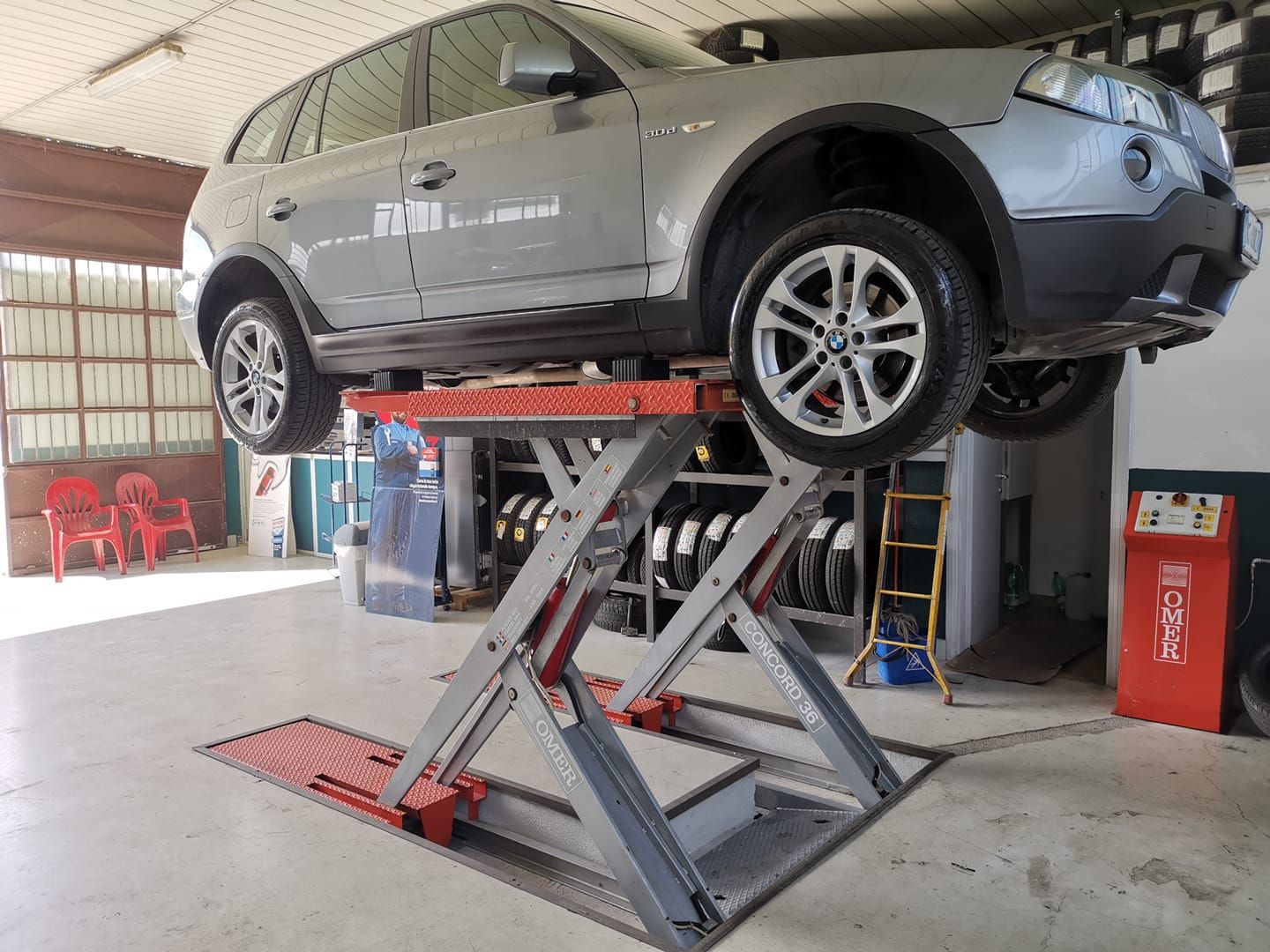 Un SUV grigio su un ponte sollevatore idraulico in un garage, pneumatici visibili, targa di sicurezza rossa.