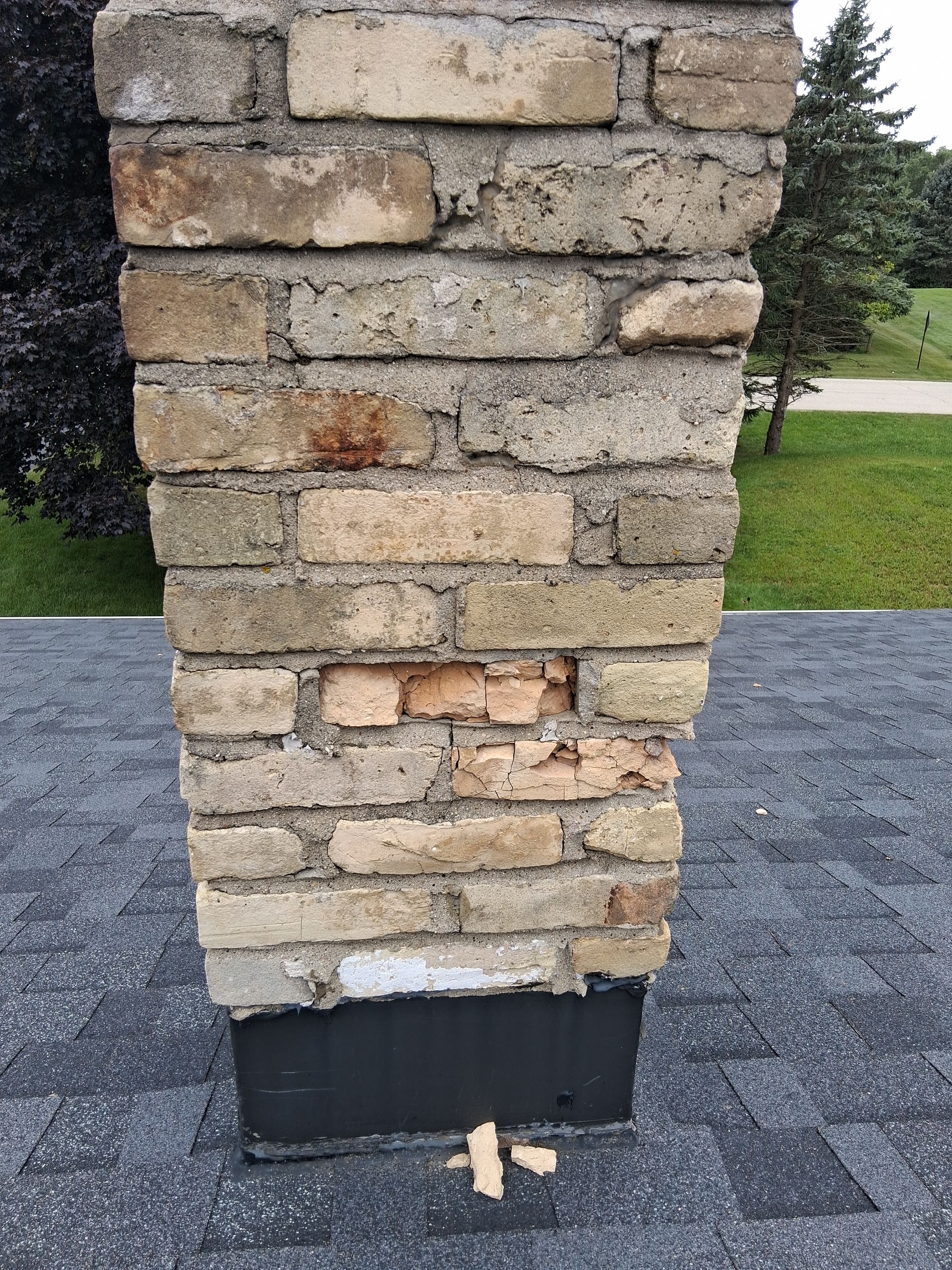 Brick chimney on a dark roof shows crumbling mortar and damaged bricks, outdoors.