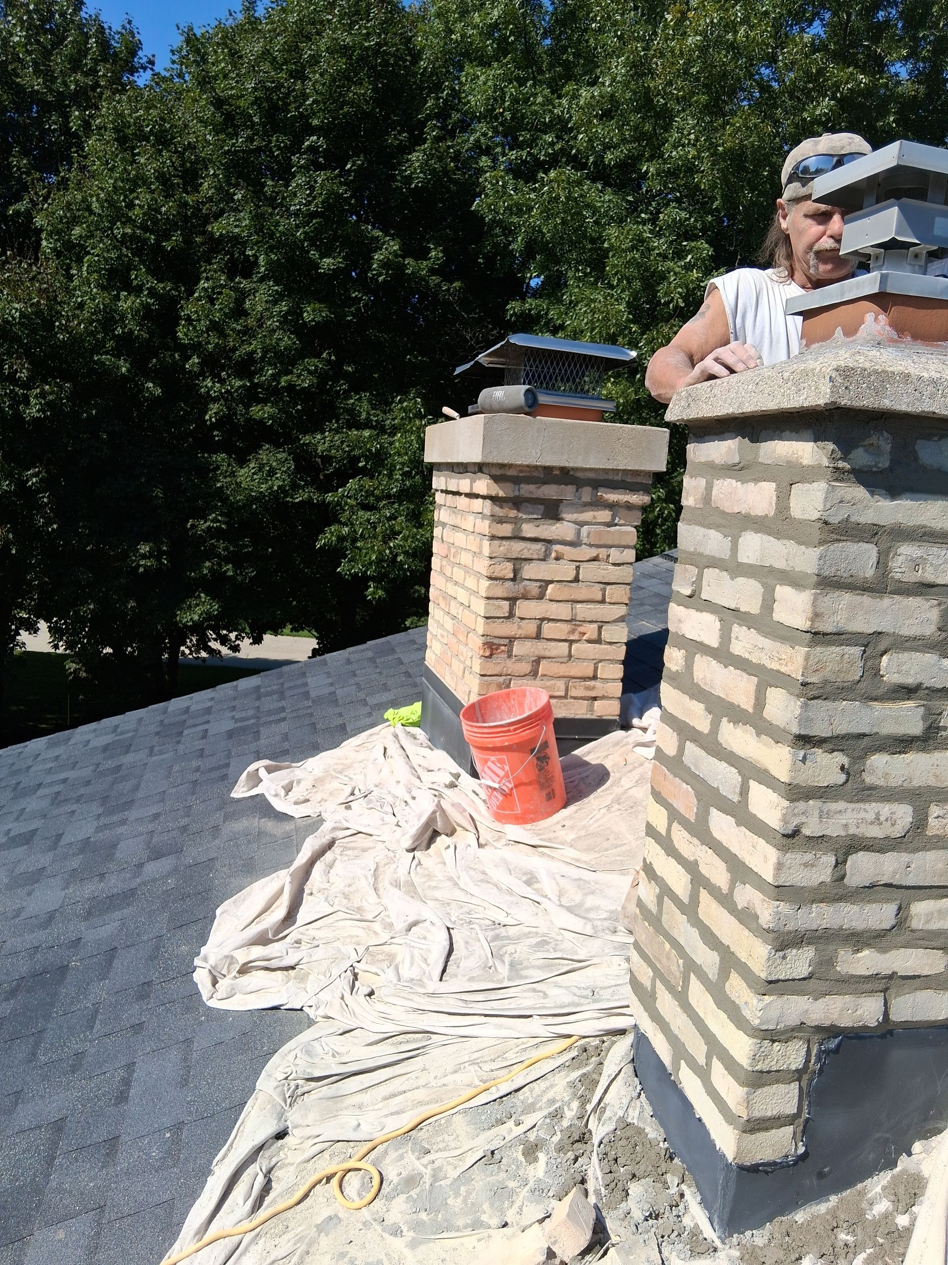 Person on a roof working on brick chimney. Red bucket and tools visible.