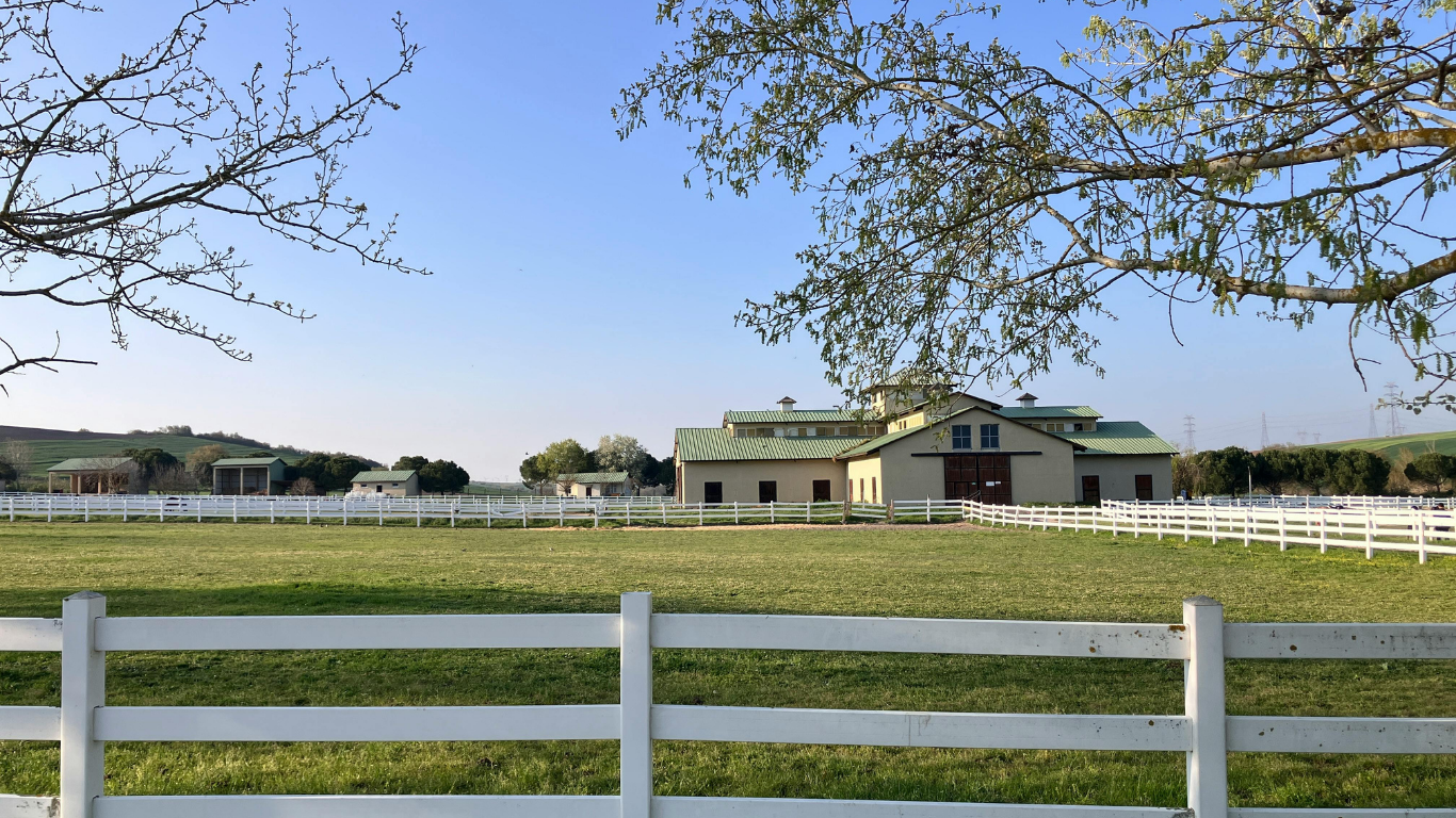 Barn with green roof, white fence, and grassy field under a blue sky.