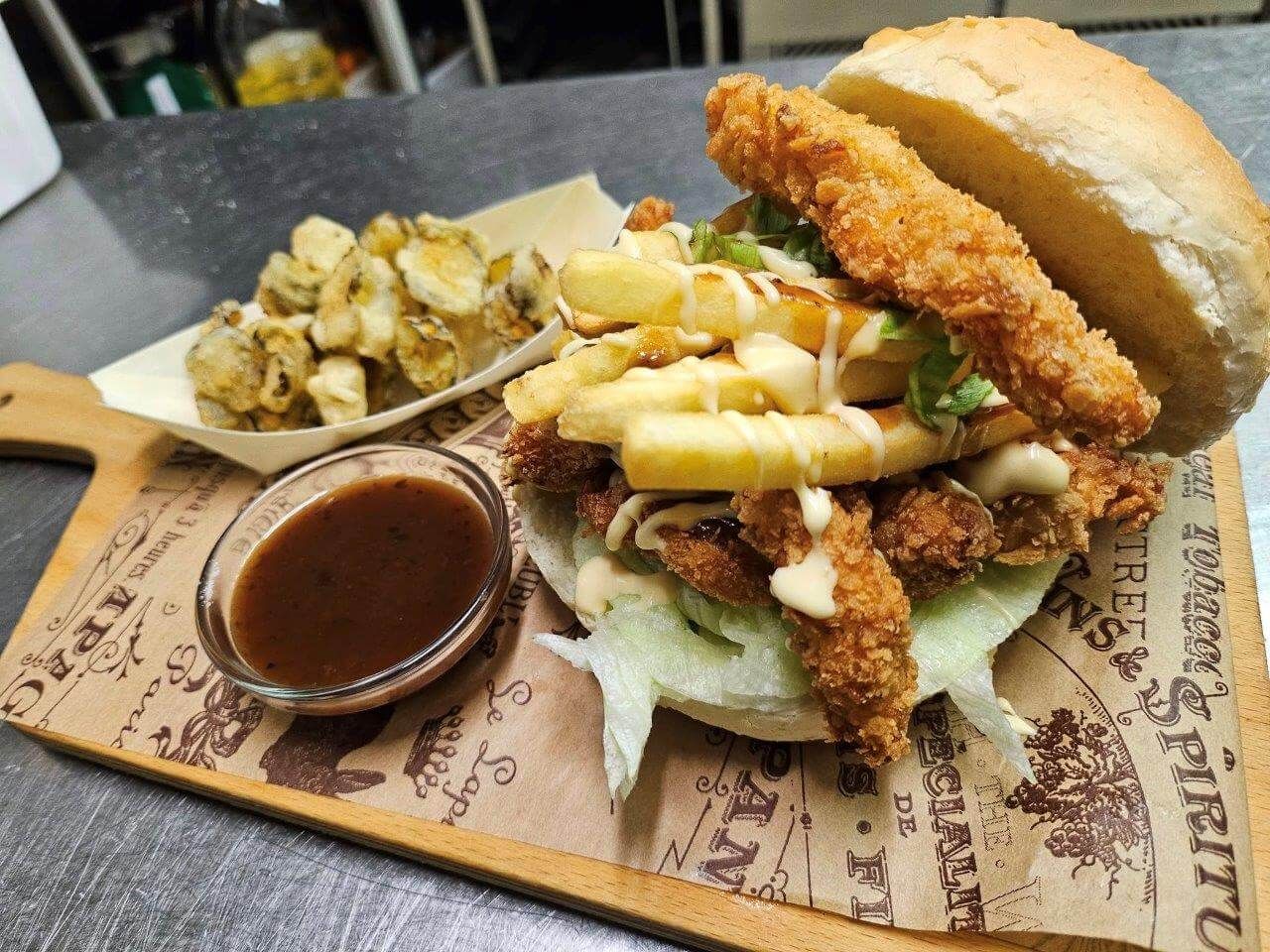 Un panino al pollo fritto con patatine fritte e salsa su un tagliere di legno.