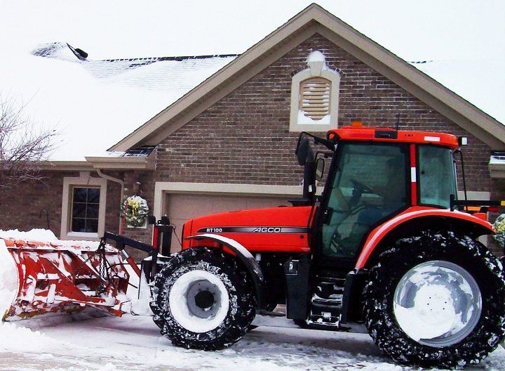 Red tractor with snowplow clearing snow in front of a brick house.