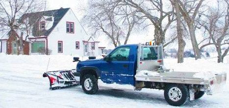 Blue pickup truck plowing snow on a snowy road with a house in the background.