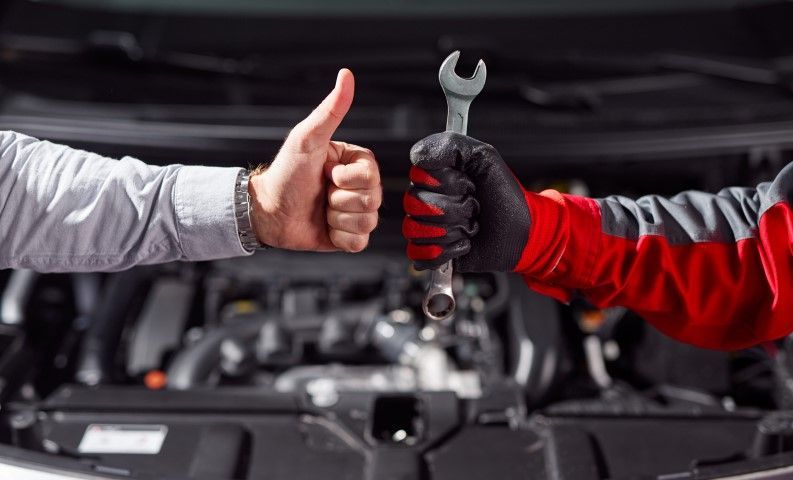 Mechanic’s hands: one gives thumbs up, the other holds a wrench over an open car engine
