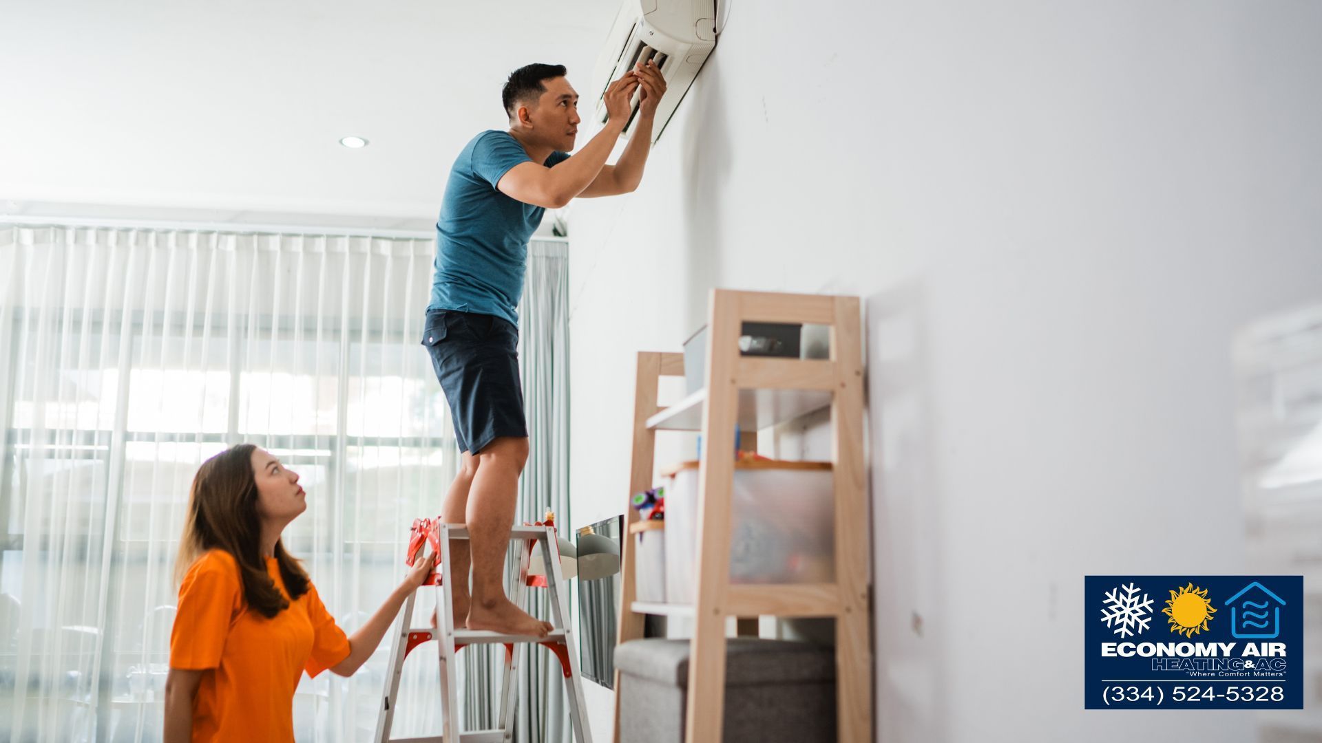 A man is standing on a ladder fixing an air conditioner while a woman looks on.