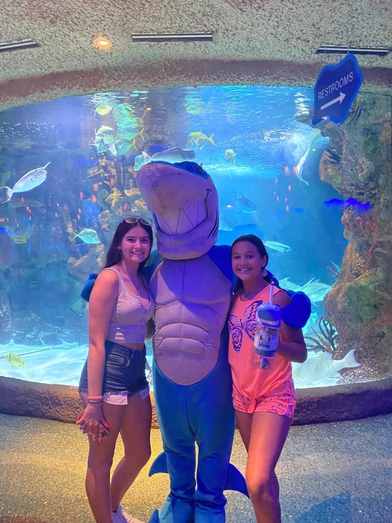 Two girls pose with a shark mascot in front of an aquarium tank filled with fish.