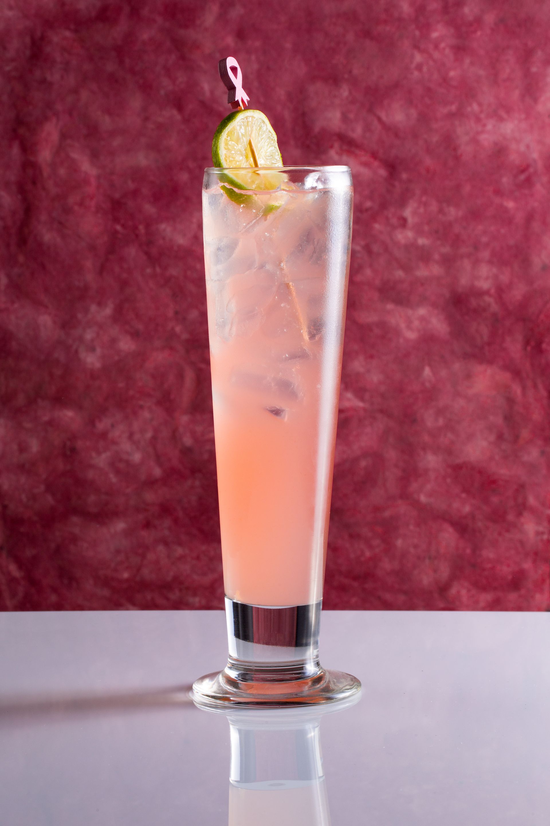 Tall pink cocktail in a glass, garnished with lime, on a white surface, red background.