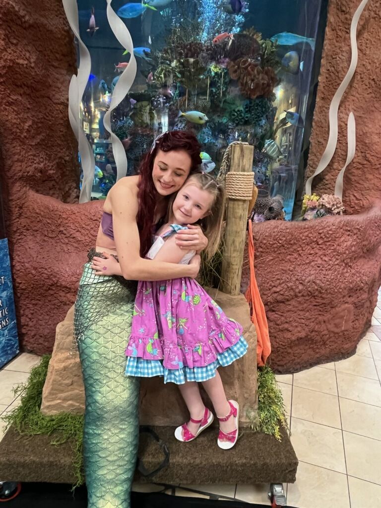 Mermaid hugs a young girl, both smiling, in front of an aquarium.