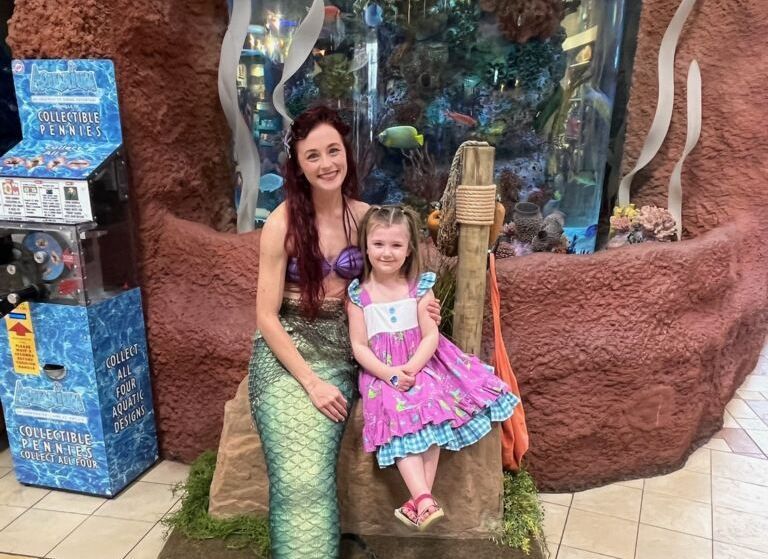 A mermaid and a young girl pose by an aquarium. The mermaid is in costume, the girl wears a pink dress.