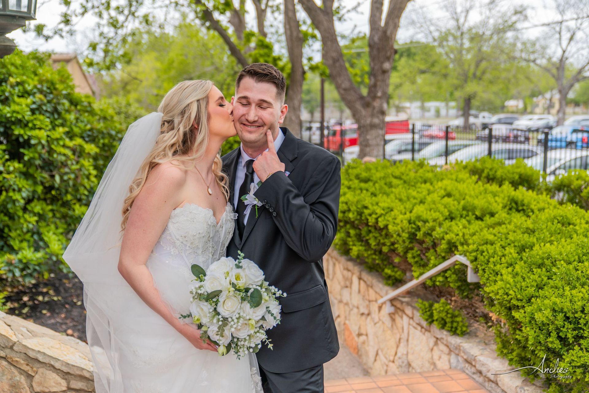 Bride and groom in wedding attire smiling outdoors near stone steps and trees