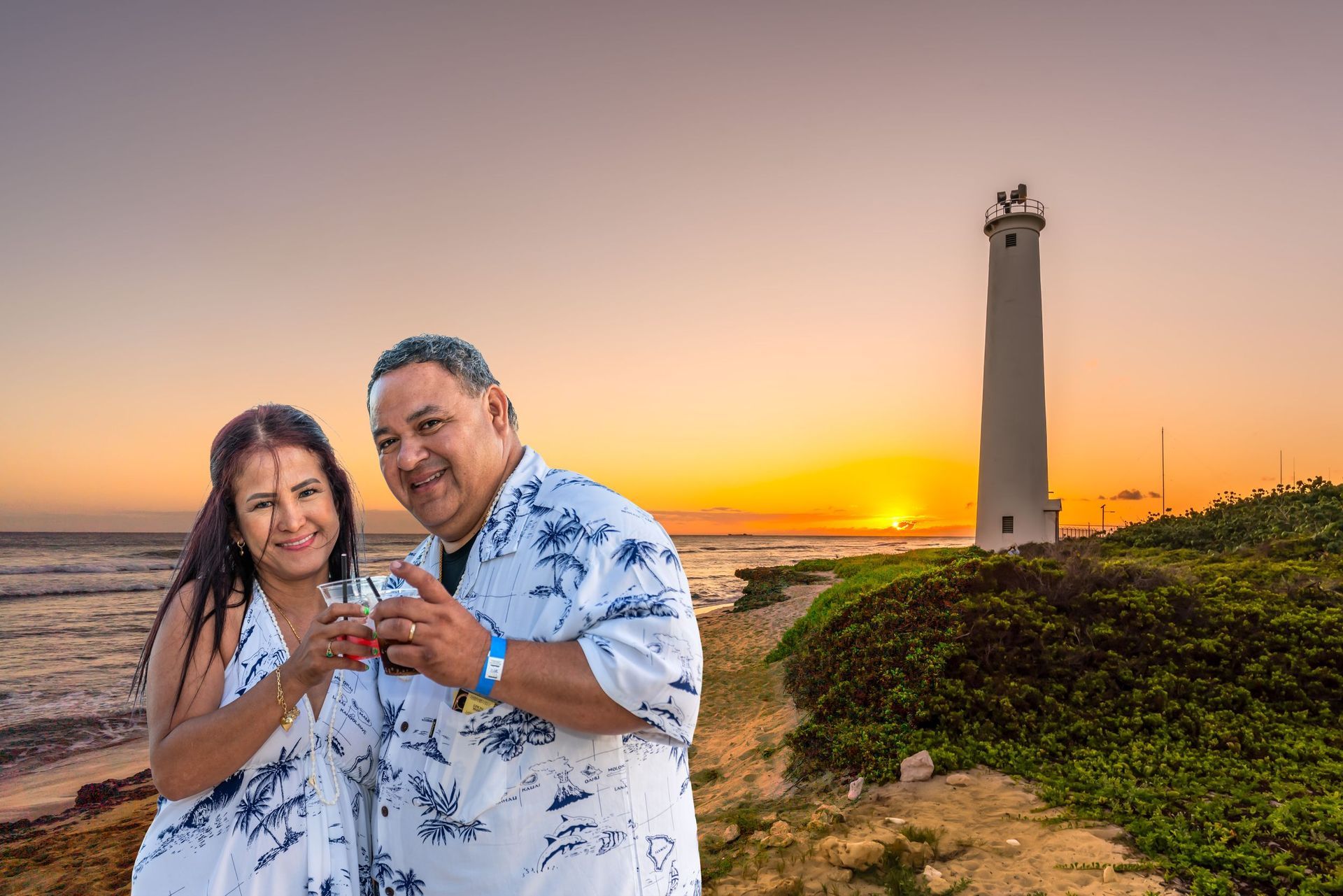 Couple holding drinks at sunset near a tall lighthouse by the coast