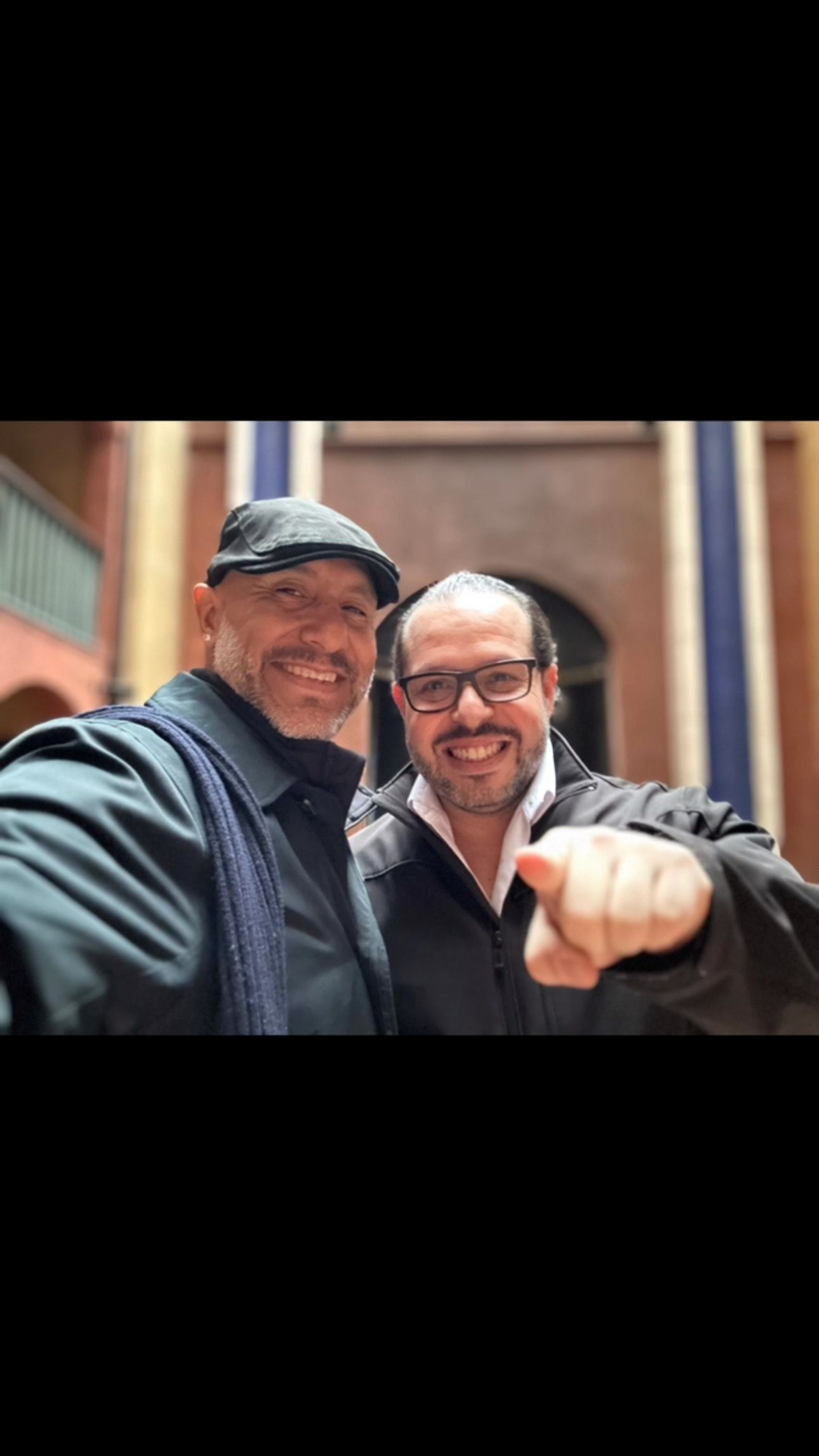 Two smiling men posing outdoors, one pointing at the camera in front of a brick building.