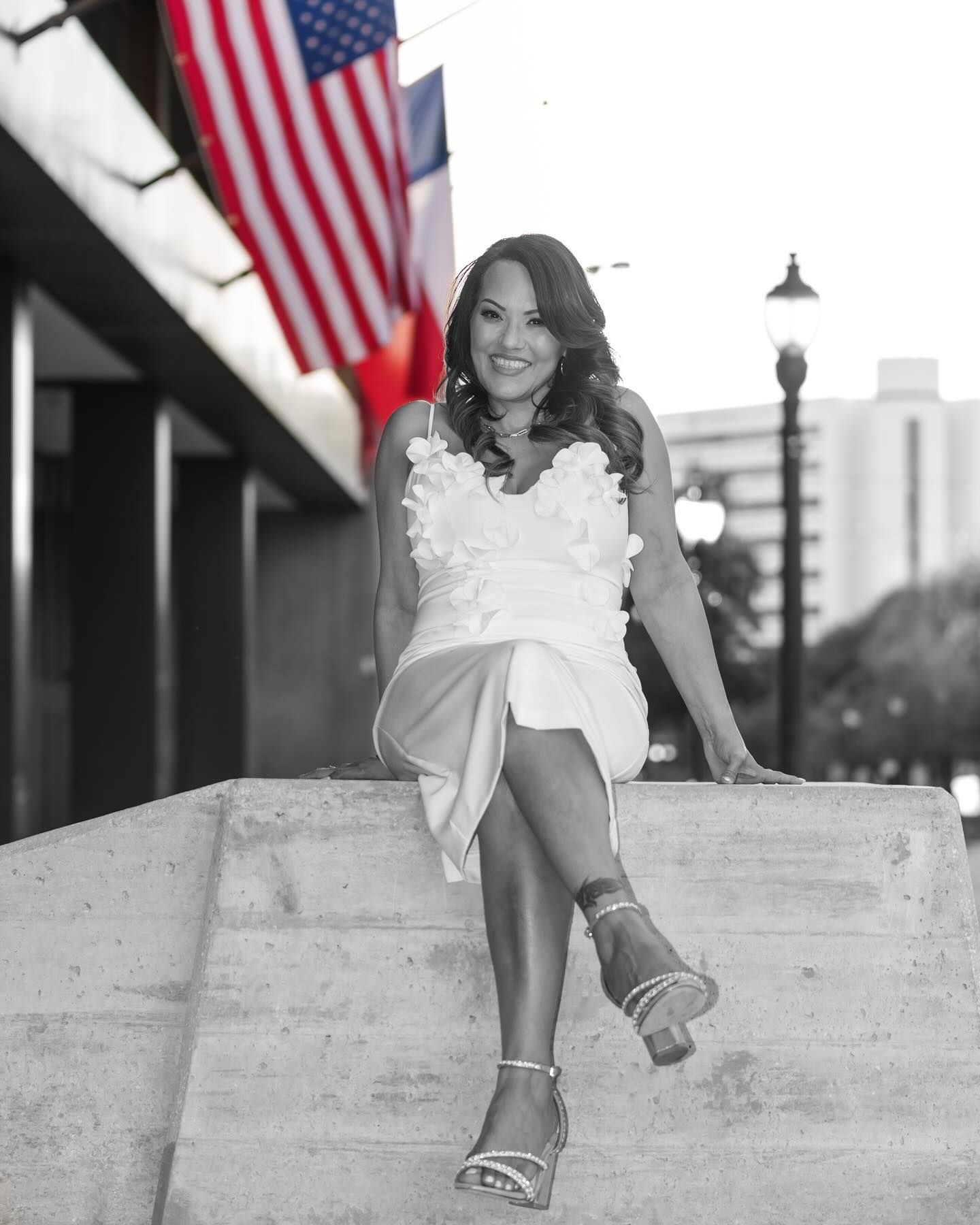 Woman in a white dress smiling on concrete steps, with an American flag blurred in the background.