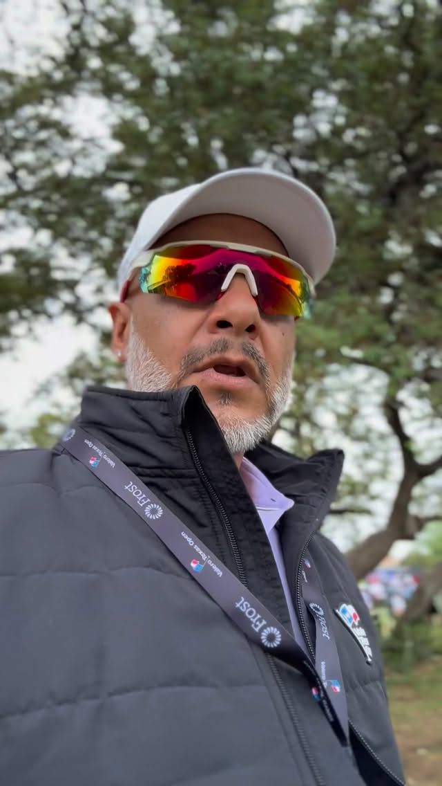 Person in a white cap and red sunglasses, wearing a black jacket outdoors with trees in the background