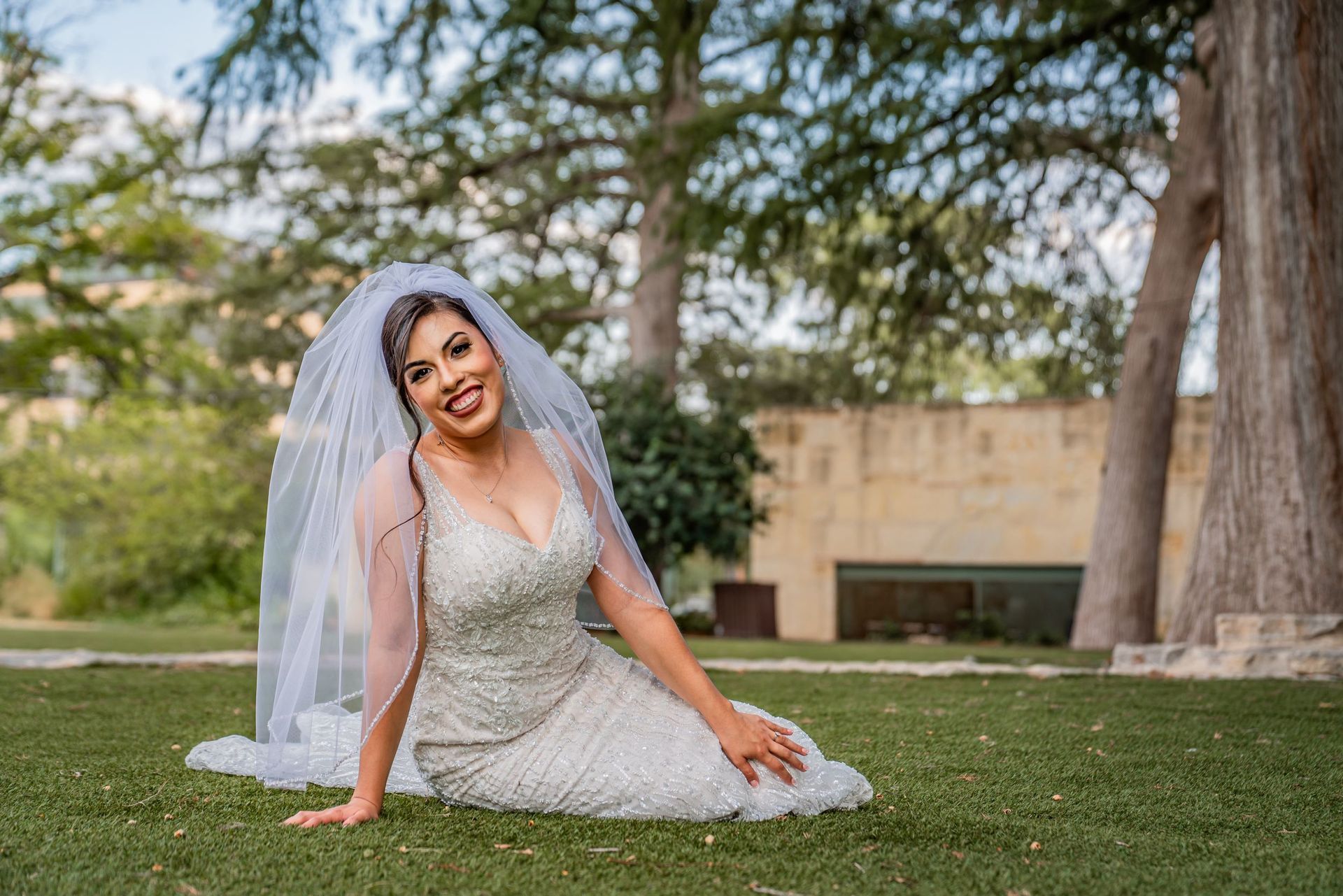 Bride in a white wedding dress and veil sitting on grass in a park, smiling at the camera.