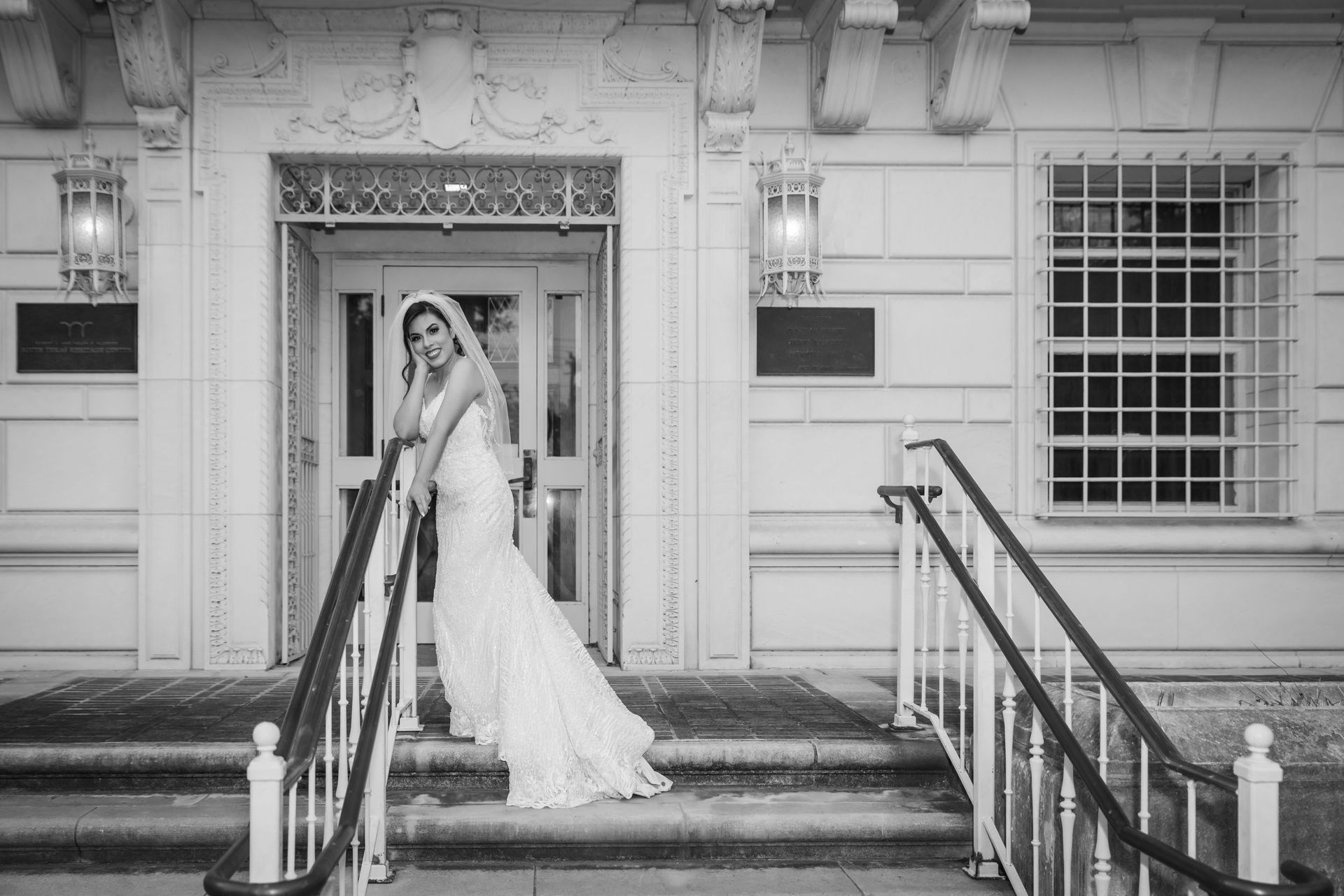 Bride in a white gown standing on steps outside a building, holding a bouquet under an ornate doorway